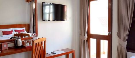 A hotel room with a bed, a desk with a kettle, a chair, a TV mounted on the wall, and a sliding glass door to the outside.