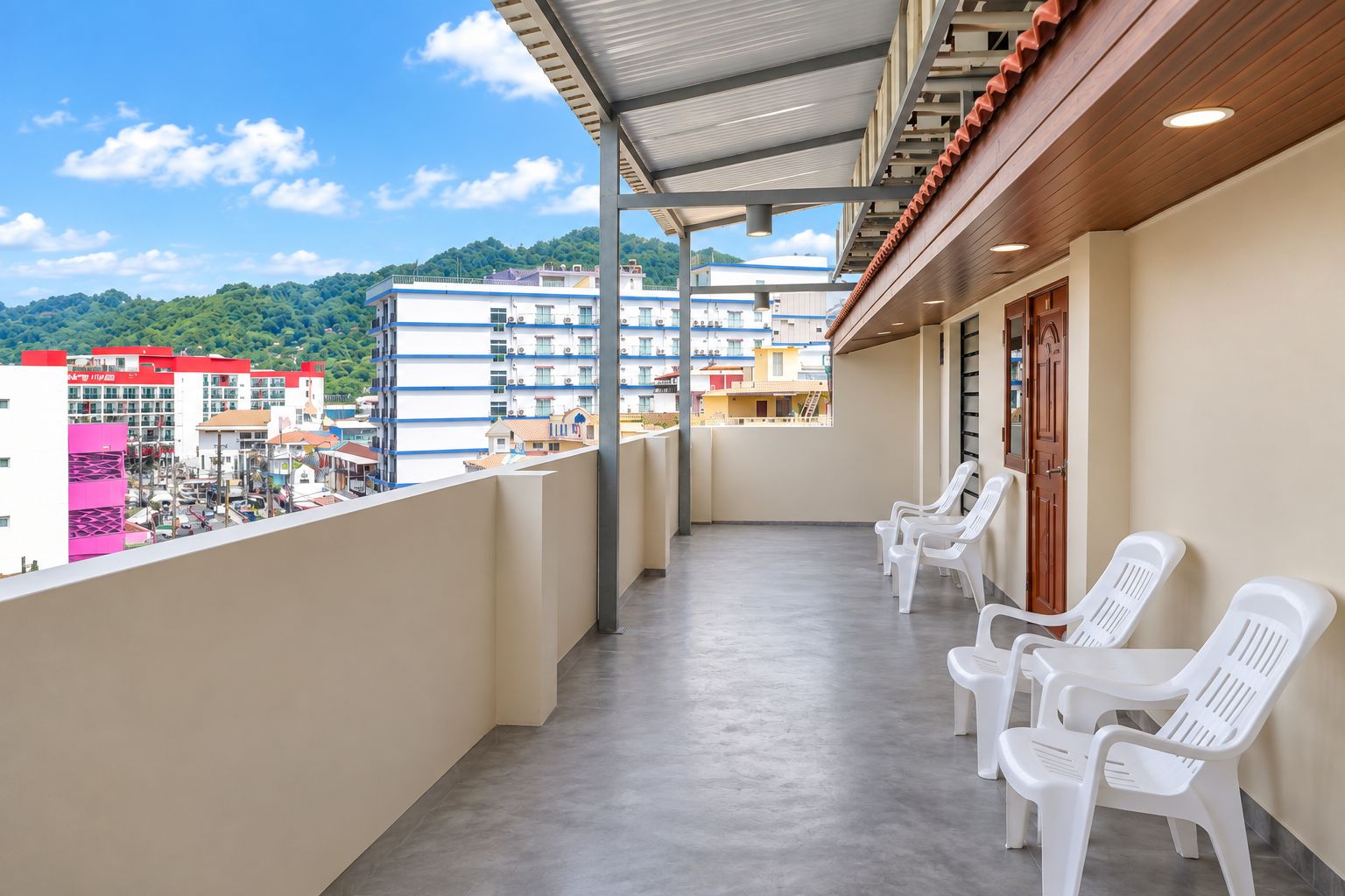 Ein Balkon mit weißen Plastikstühlen, einem Betonboden und Blick auf Gebäude und Hügel im Hintergrund.