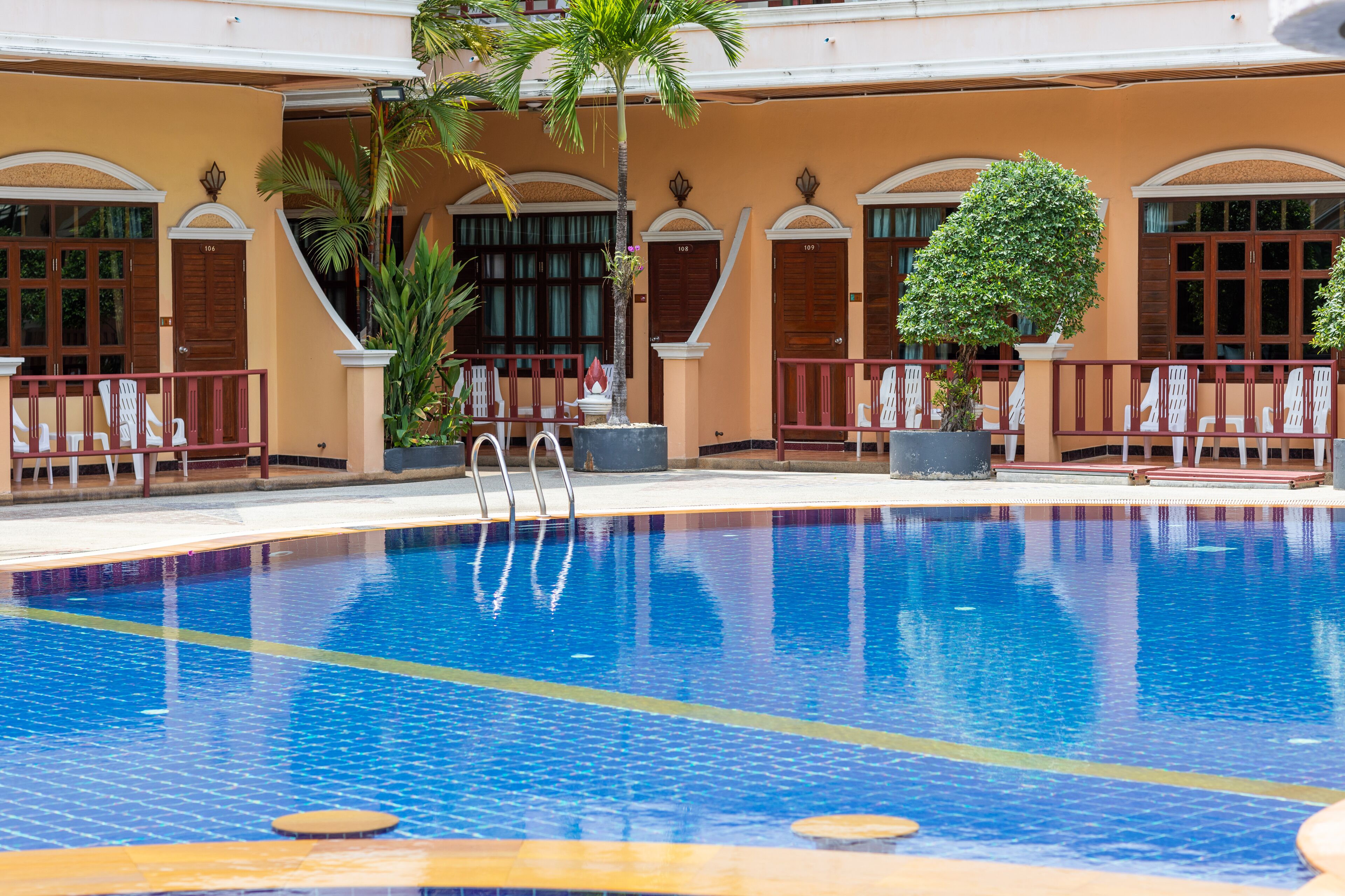 A swimming pool with a tiled deck, surrounded by a building with arched windows and doors, and potted plants.