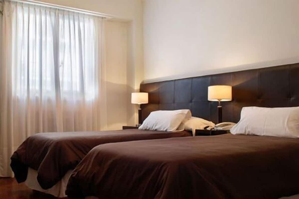 Habitación de hotel con dos camas, un ventanal con cortinas translúcidas y un cabecero oscuro.