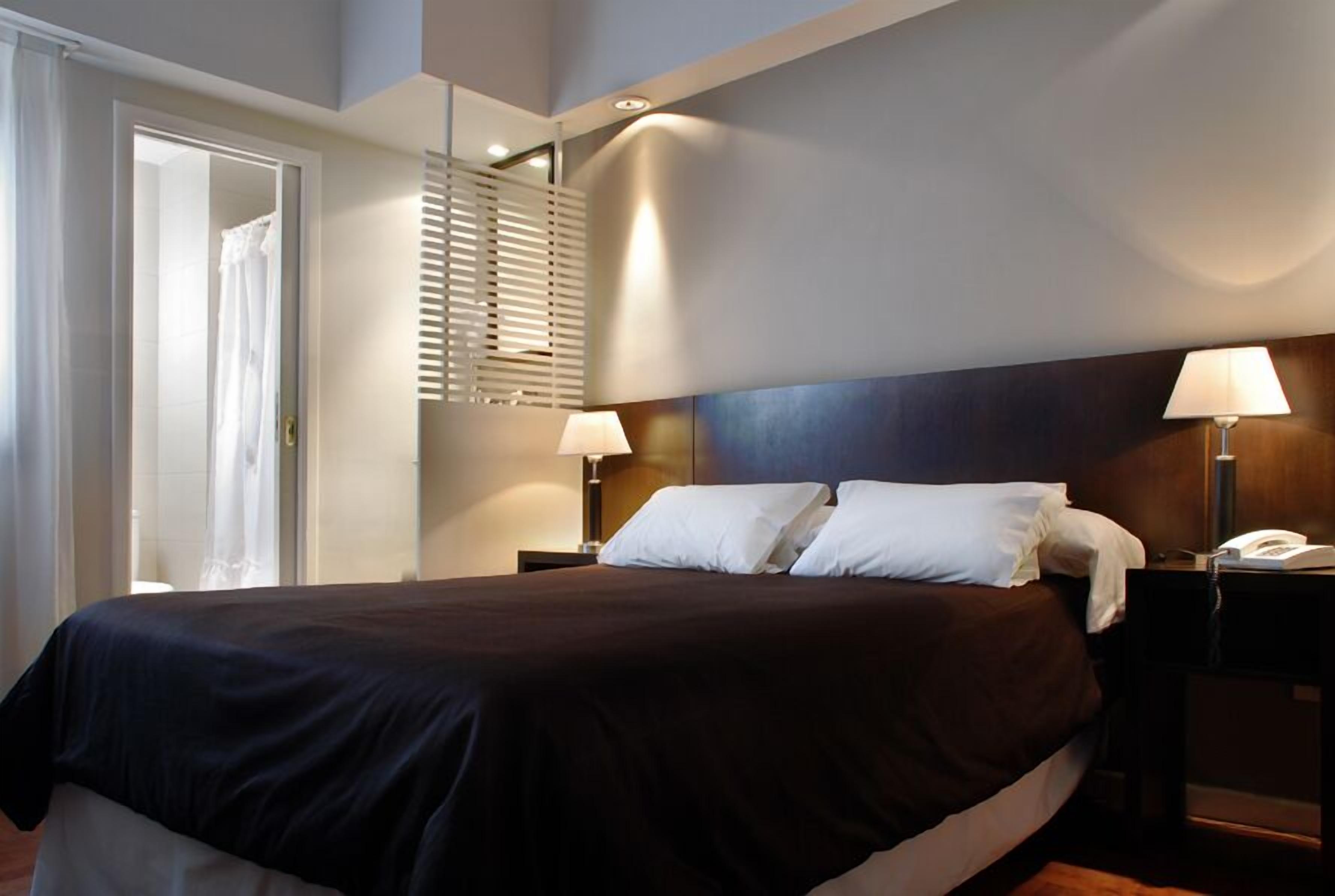 A hotel room with a large bed, two bedside lamps, a wooden headboard, and a door leading to a bathroom.