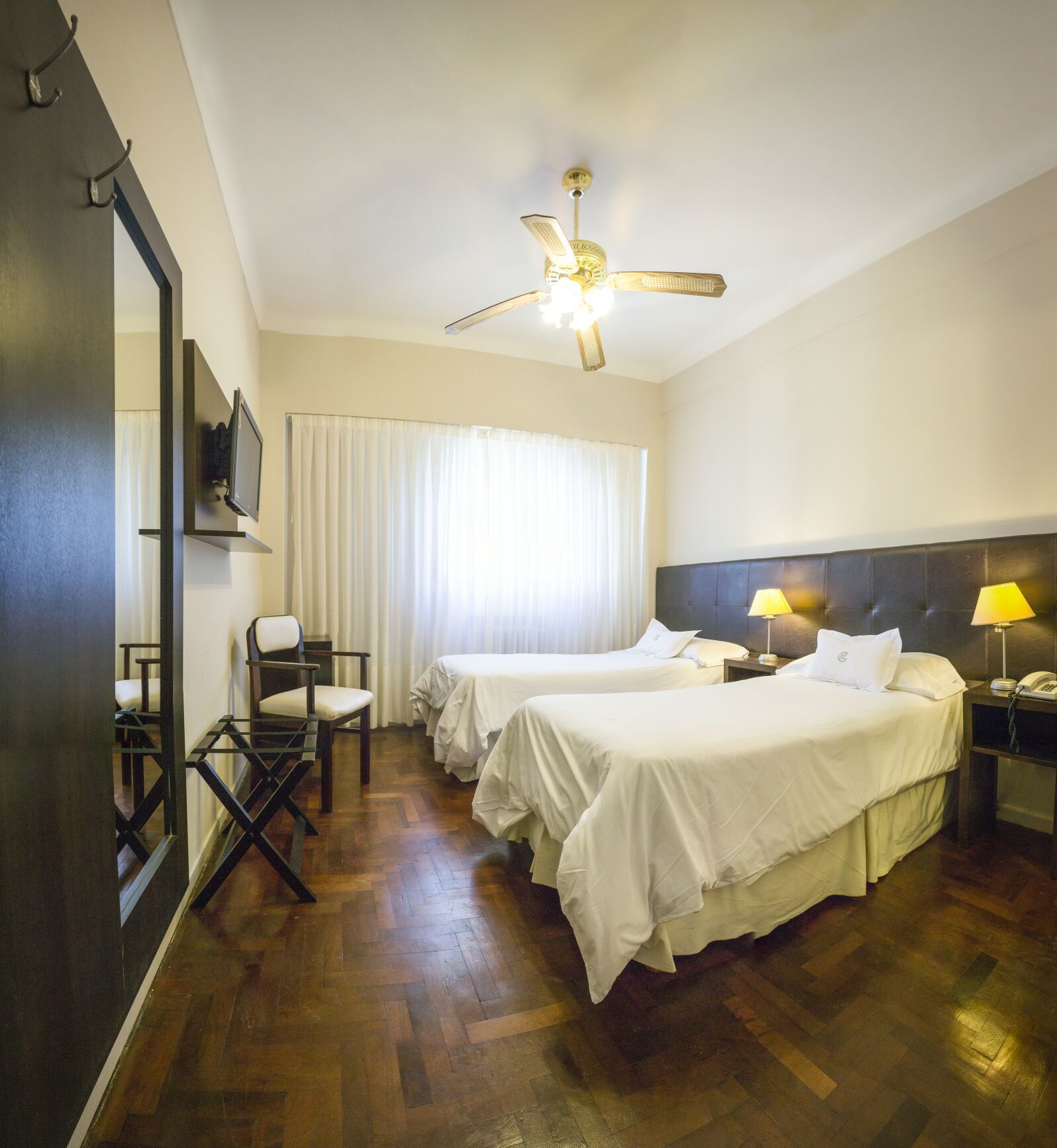 A hotel room with two beds, a ceiling fan, a desk with a chair, and a large mirror.
