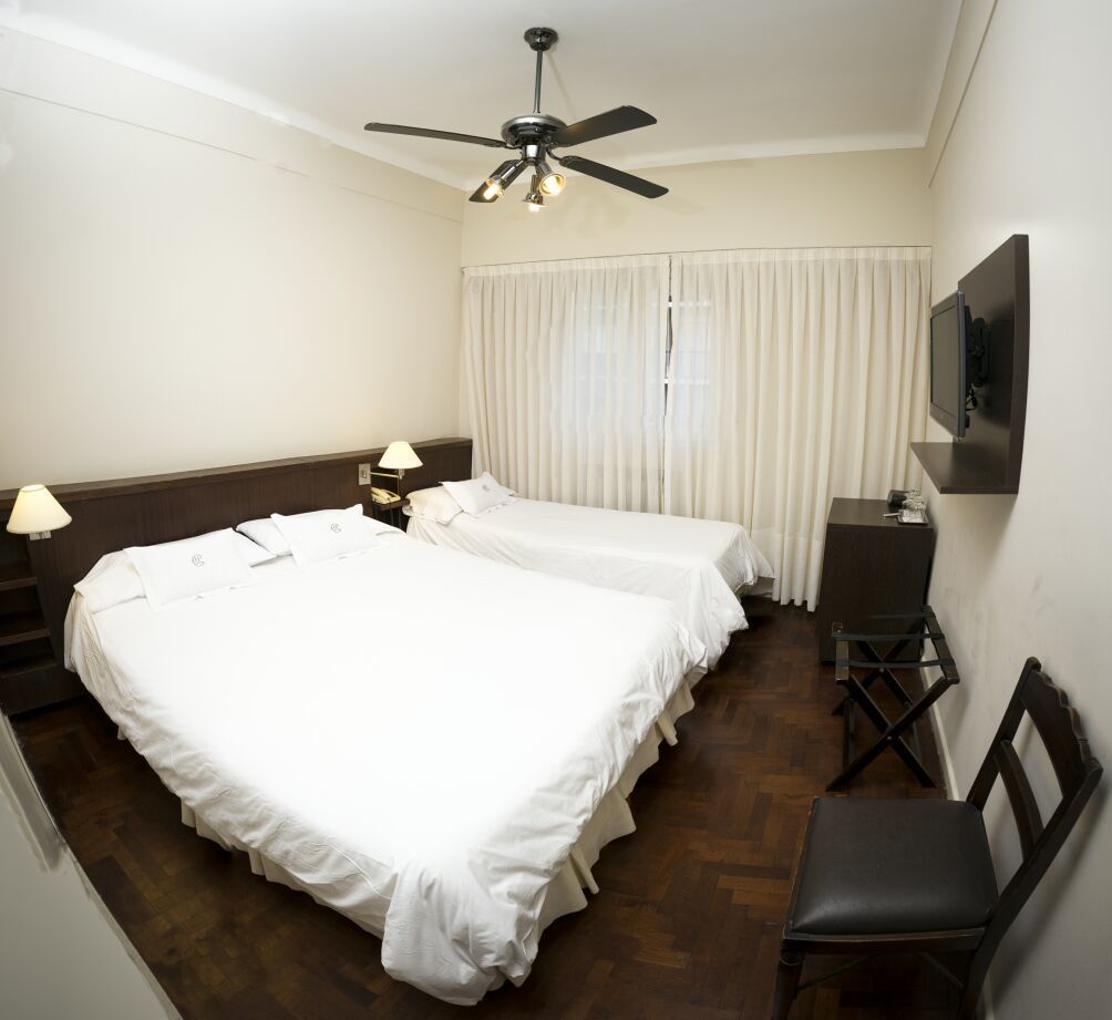 Une chambre d’hôtel avec deux lits simples, un ventilateur de plafond, un téléviseur fixé au mur, un petit bureau et une chaise.
