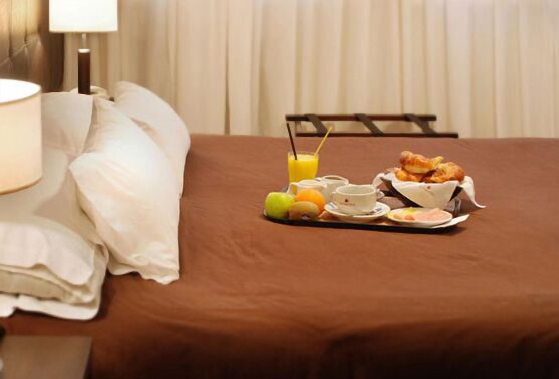 A neatly made bed with white linens and pillows, a tray of breakfast including croissants, fruit, and orange juice, and a lamp on the side.