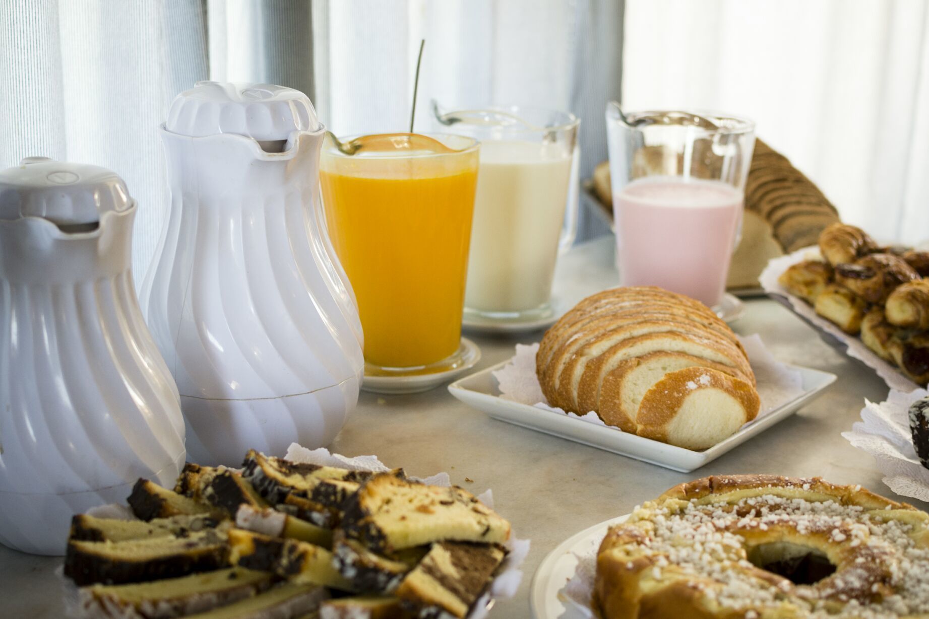 Un desayuno con diversas opciones de pastelería, un croissant y tres bebidas en jarras y vasos.