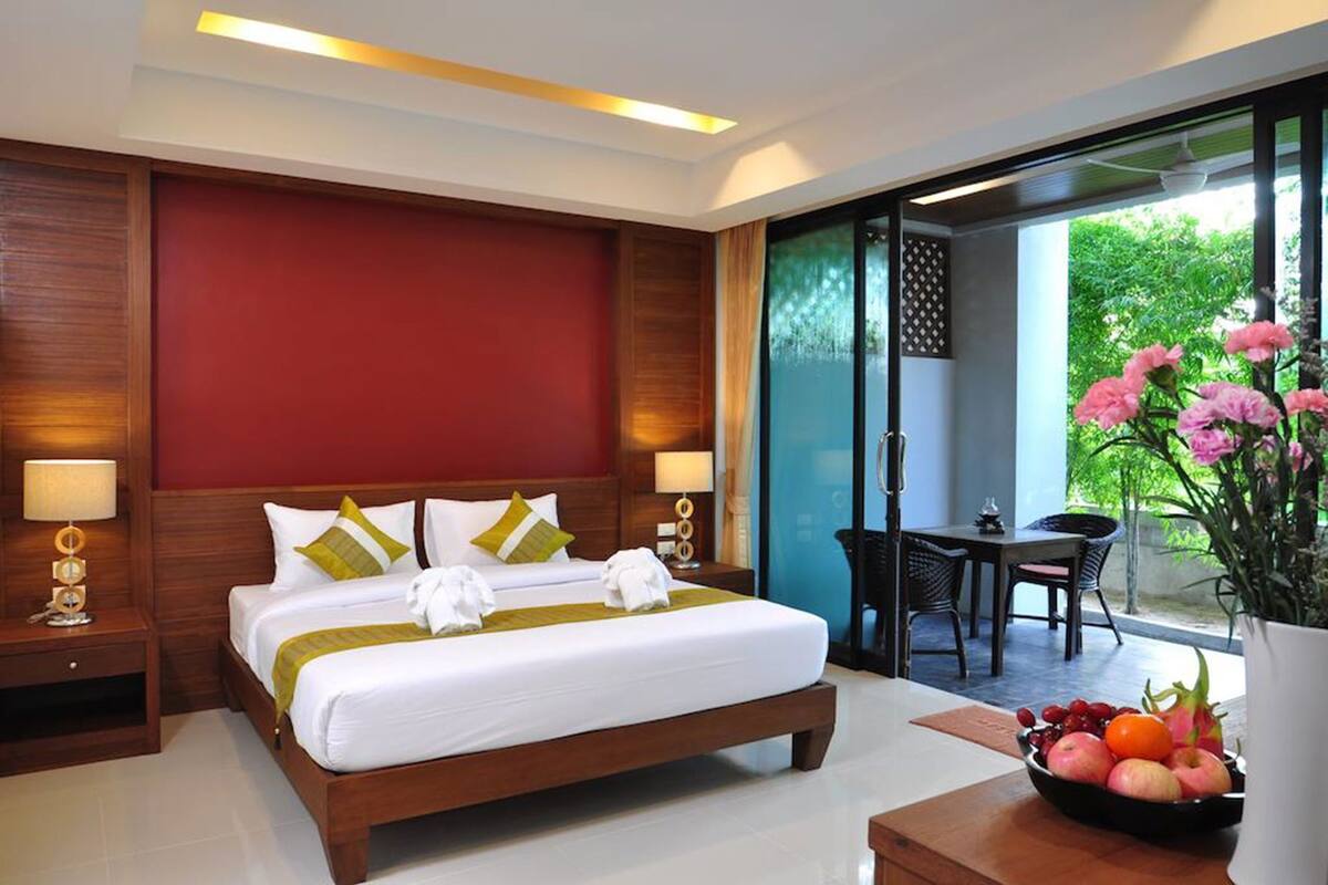 Quarto de hotel com uma cama grande, duas mesas de cabeceira, uma escrivaninha com cadeira, uma mesa de jantar com cadeiras e uma varanda florida.