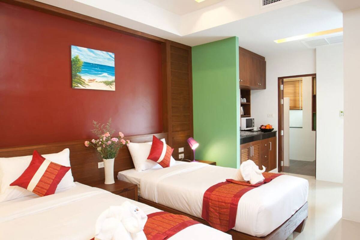 Quarto de hotel com duas camas, um quadro na parede, uma parede de destaque verde e uma kitchenette ao fundo.