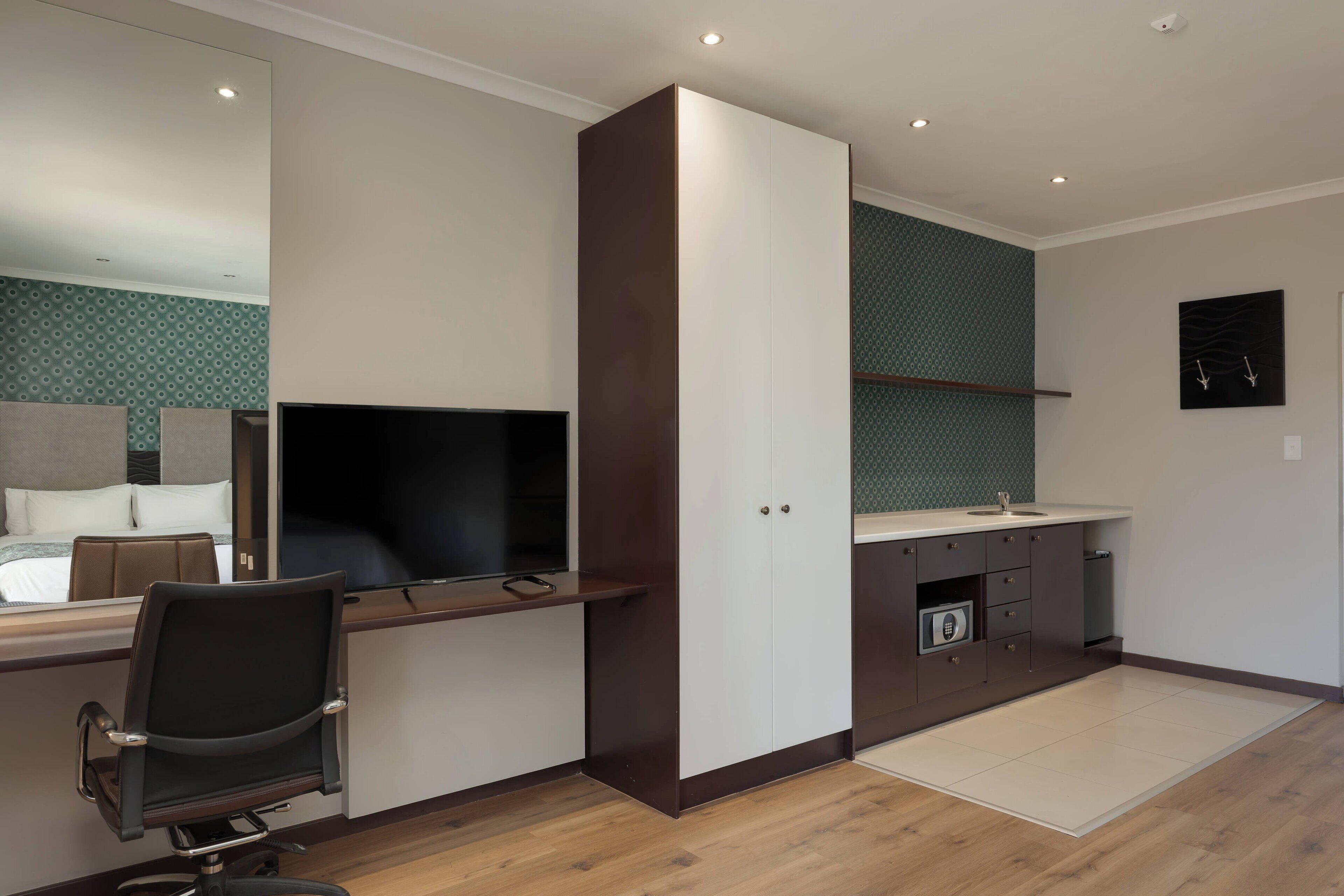 A modern hotel room with a bed, a desk with a chair, a TV, a wardrobe, a bathroom with a sink and mirror, and a wall-mounted artwork.