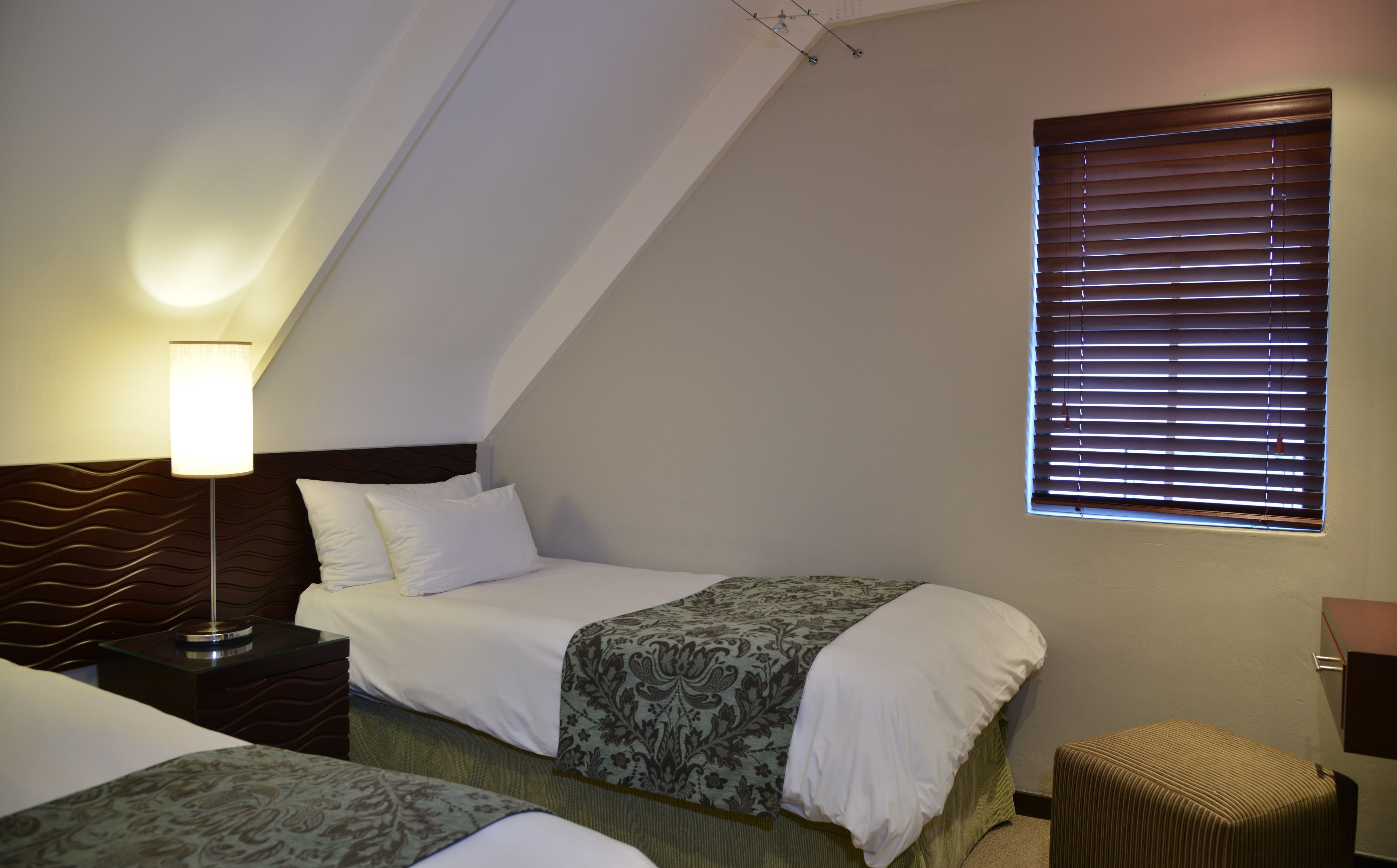 Ein Schlafzimmer mit zwei Betten, einem Nachttisch, einer Lampe, einem Fenster mit Jalousien und einem kleinen Hocker.