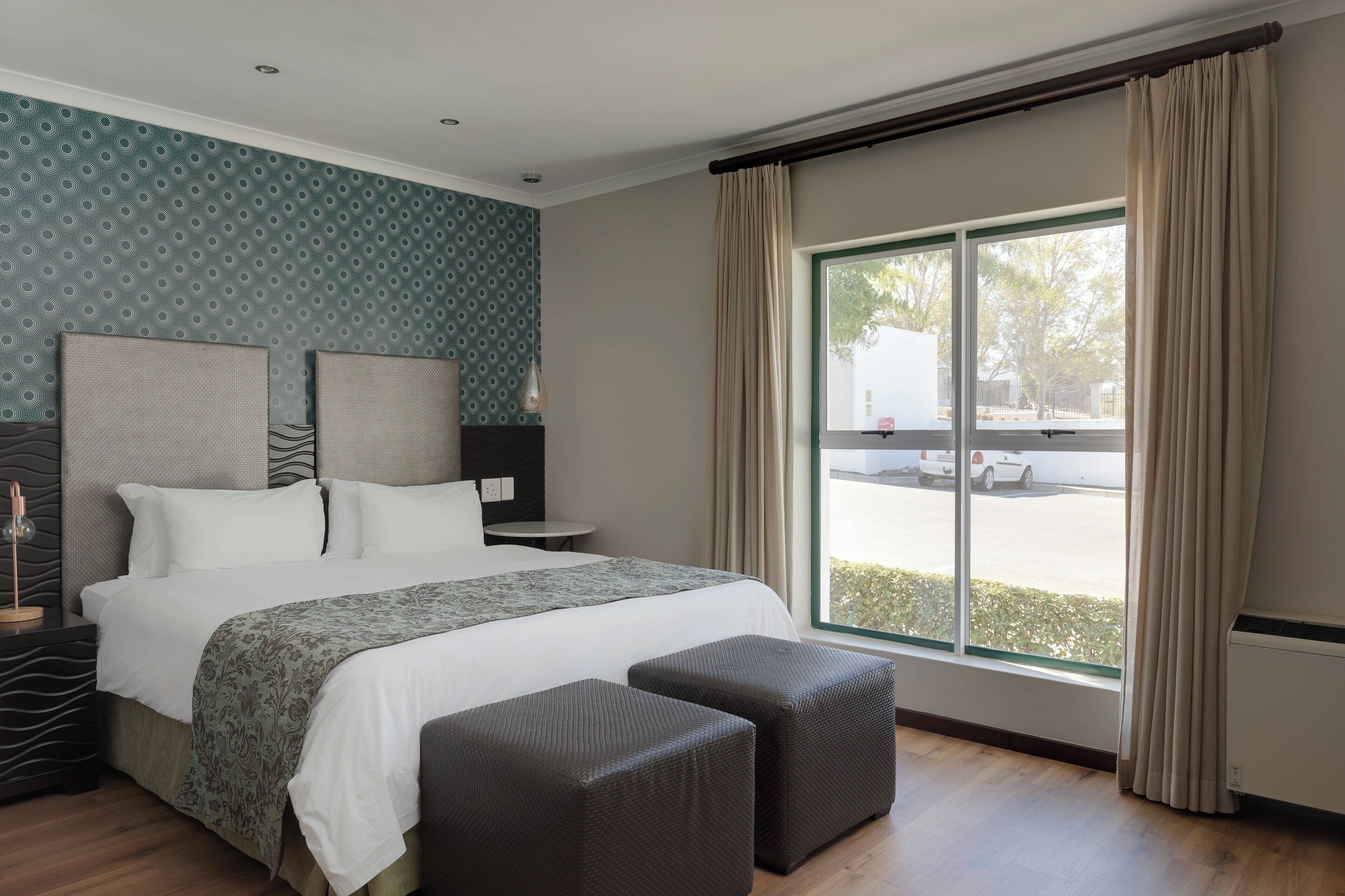 A modern hotel room with a large bed, two bedside tables, a small table, and a large window with curtains.