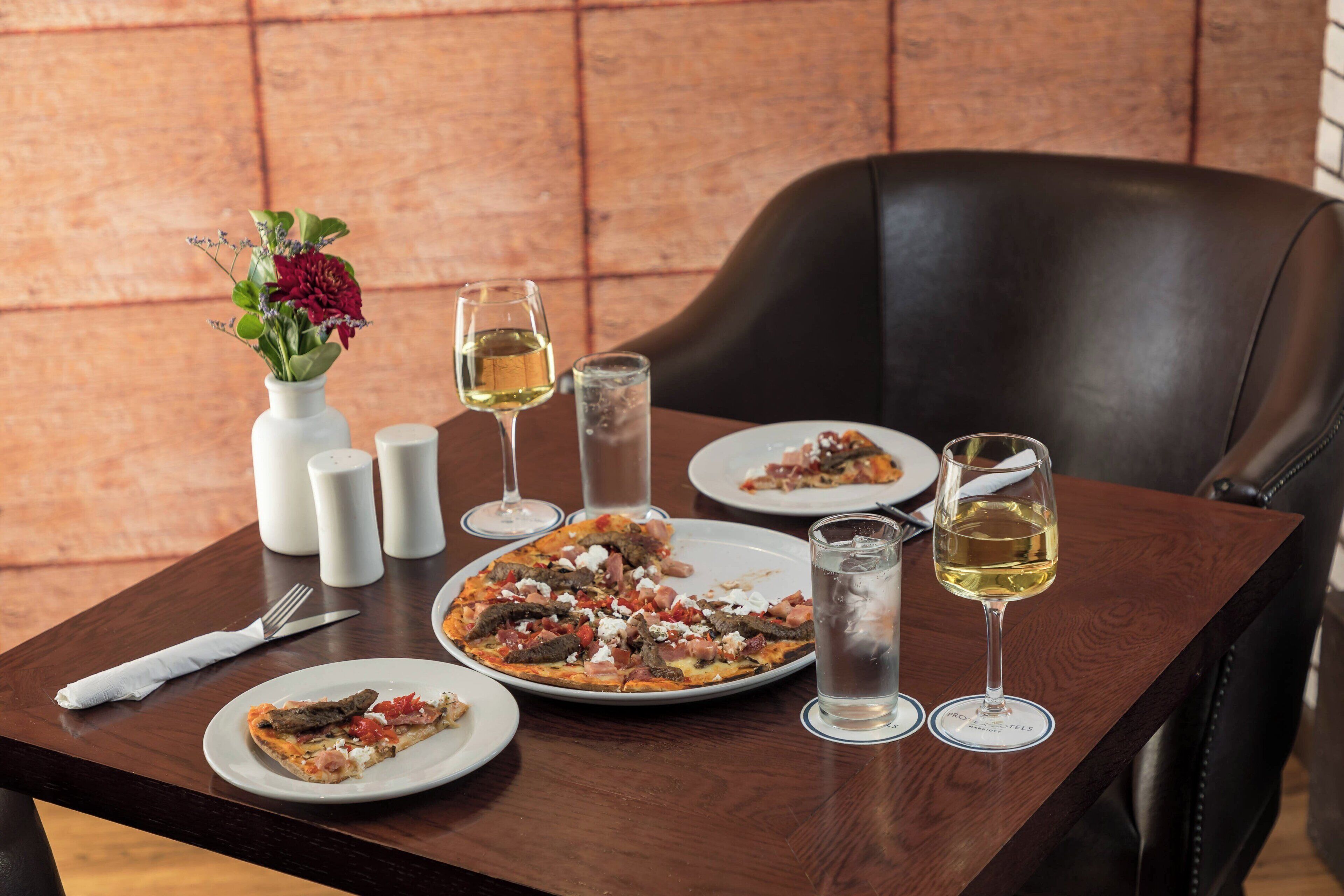Une table à manger avec une pizza, des verres à vin et un vase de fleurs.