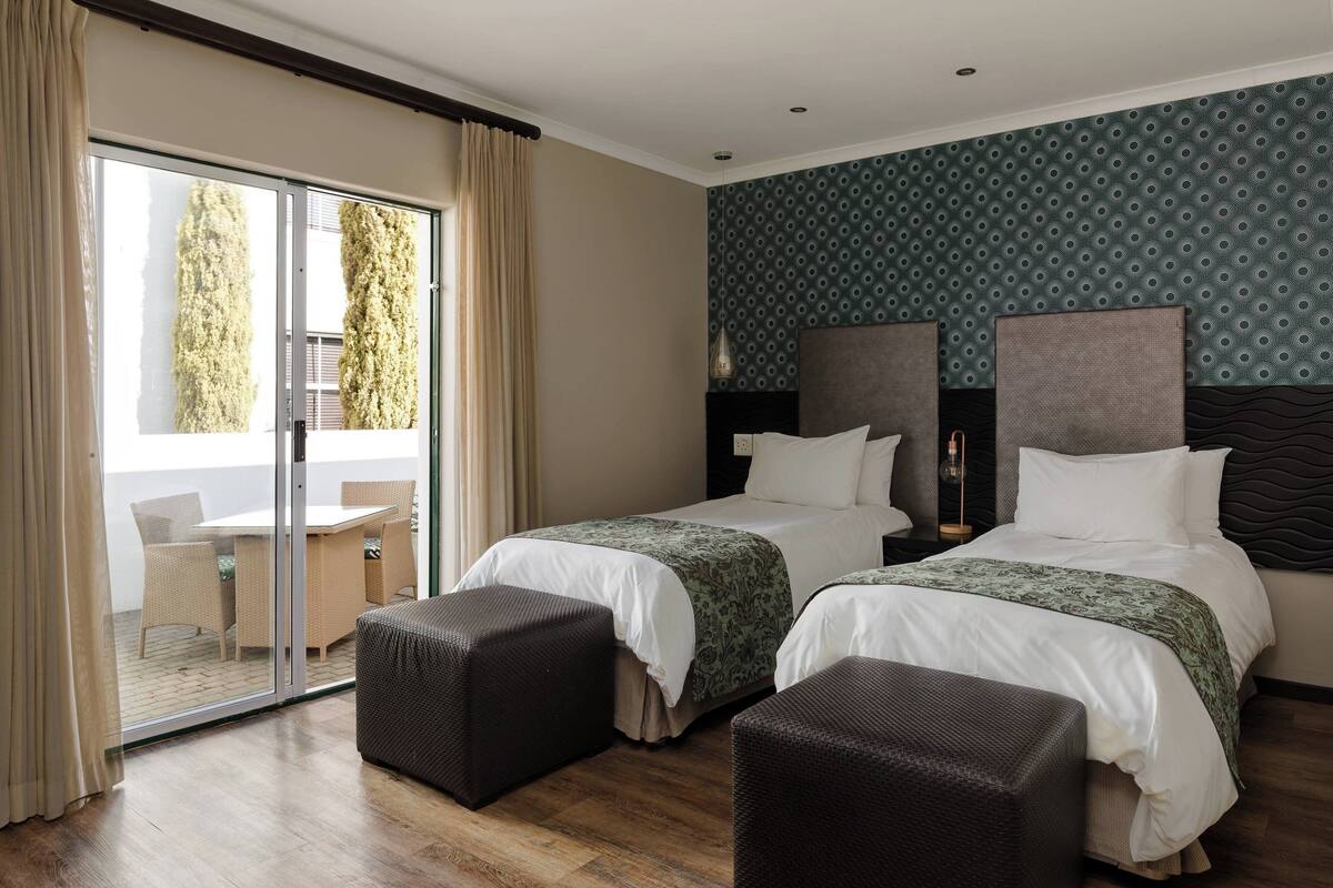 Ein Hotelzimmer mit zwei Betten, einem Balkon mit Tisch und Stühlen und Blick auf Bäume.