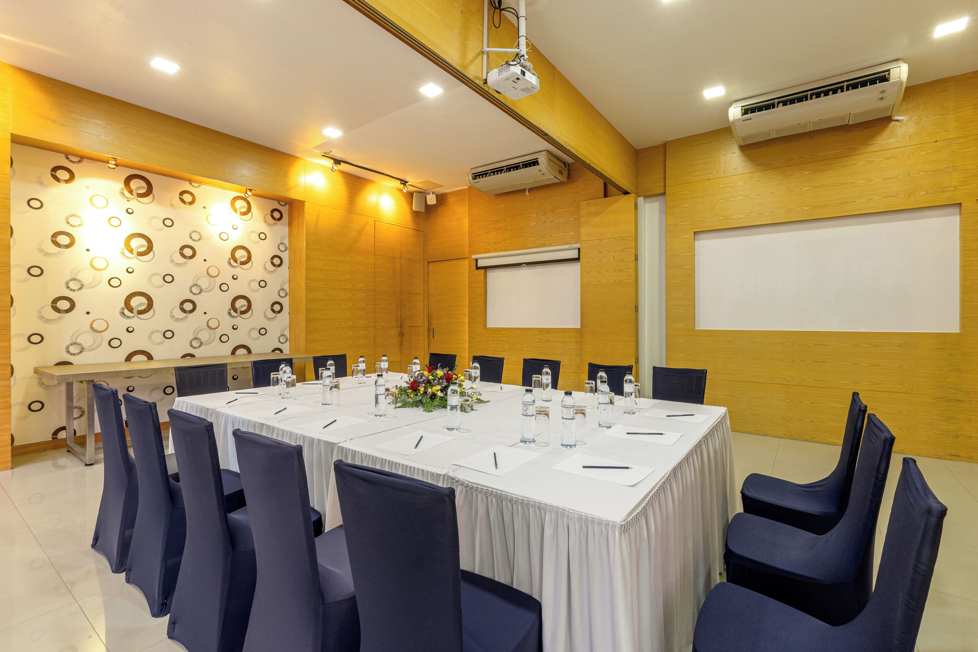 Un salón de conferencias con una larga mesa, sillas, un proyector, un equipo de aire acondicionado y una pizarra.