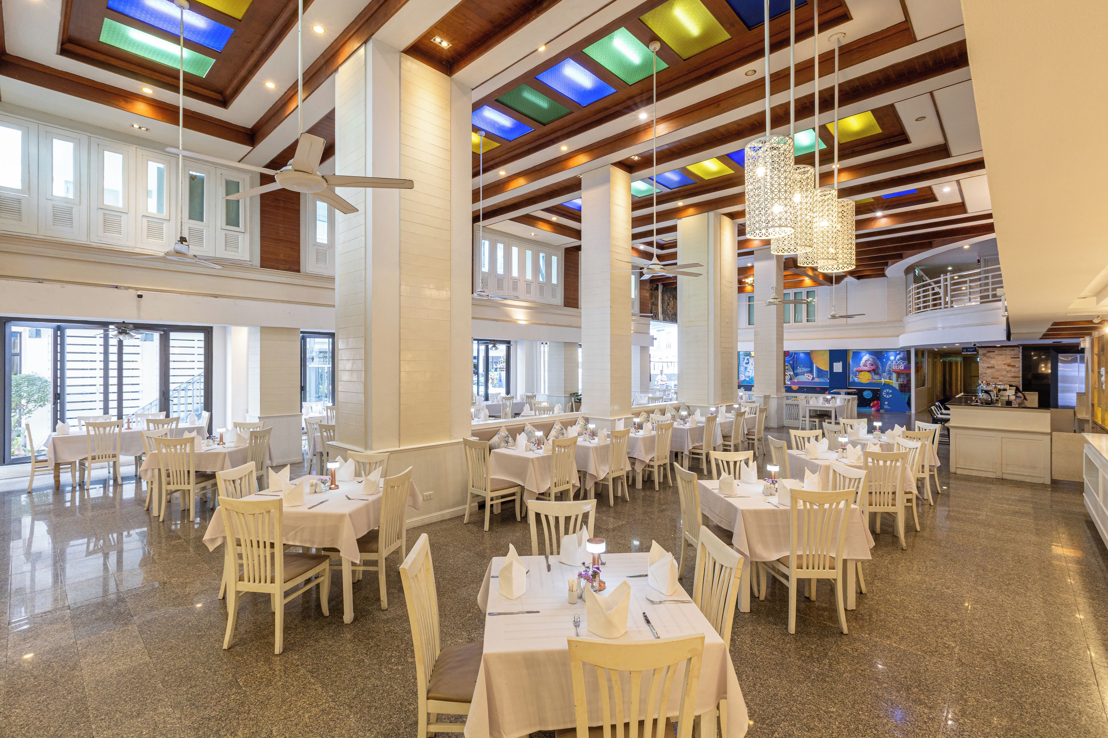 A spacious dining area with multiple tables and chairs, high ceilings with wooden beams, and large windows allowing natural light.