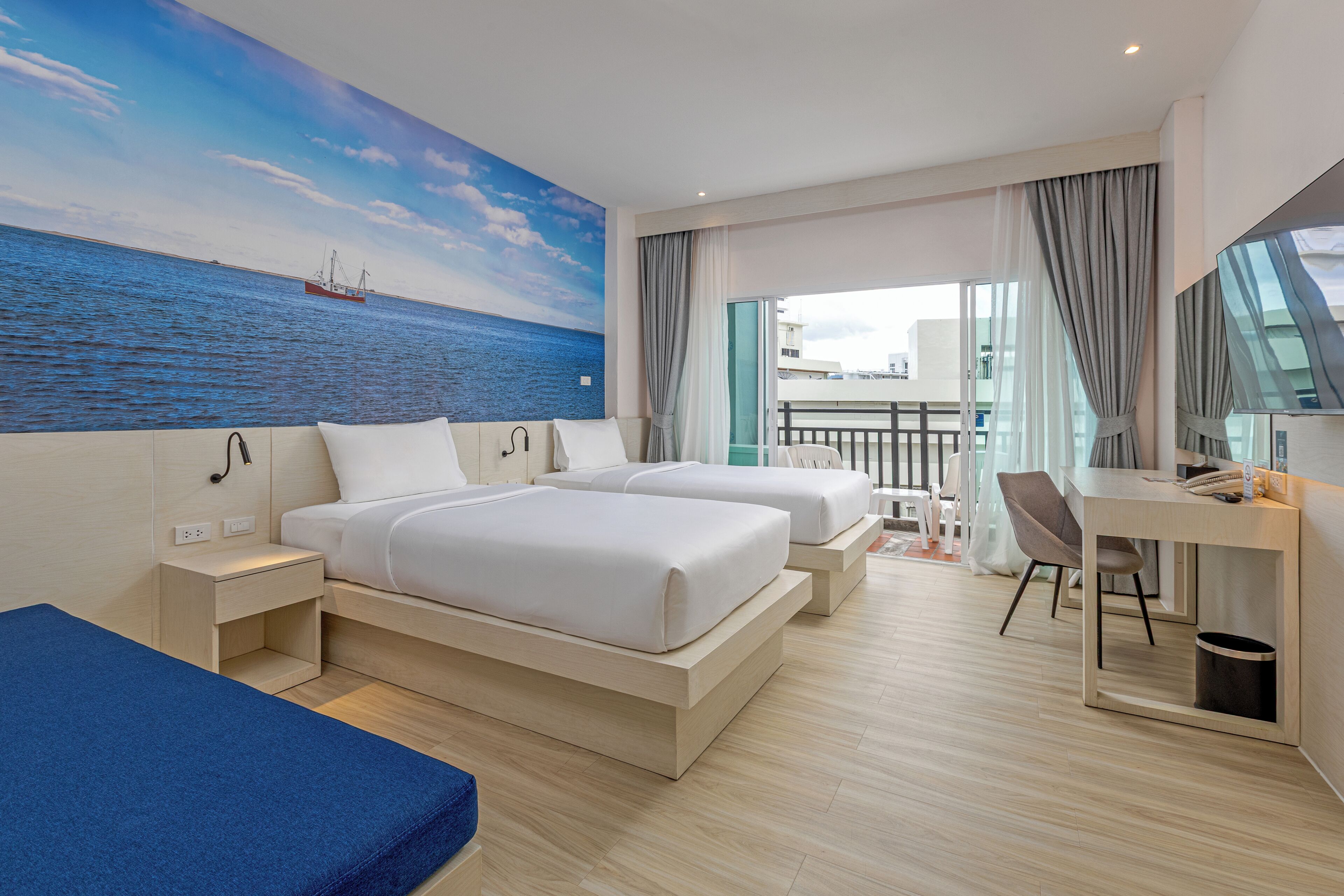 A hotel room with two beds, a desk, a chair, a balcony with a view of the sea, and a mural of the sea.