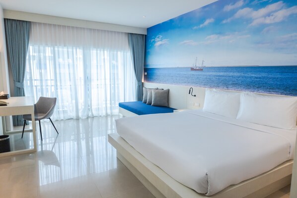A modern hotel room with a large bed, a desk, and a view of the sea through a window. - Amata Patong (Patong)