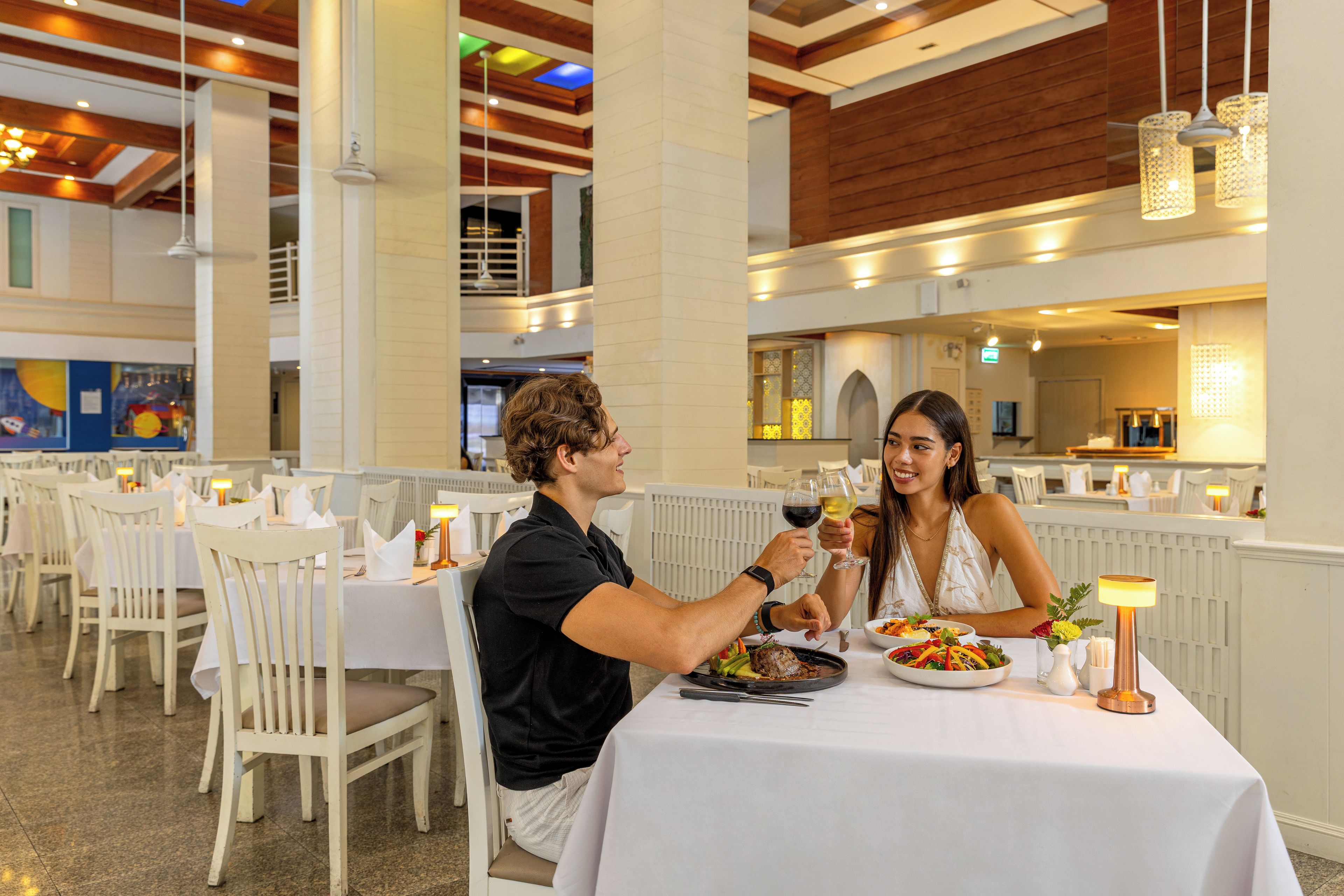 A restaurant with white tables and chairs, a high ceiling with wooden beams, and pendant lighting.