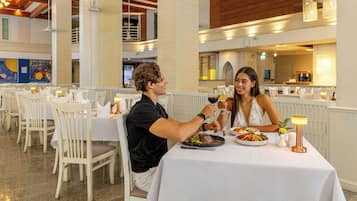 A restaurant with white tables and chairs, a high ceiling with wooden beams, and pendant lighting.