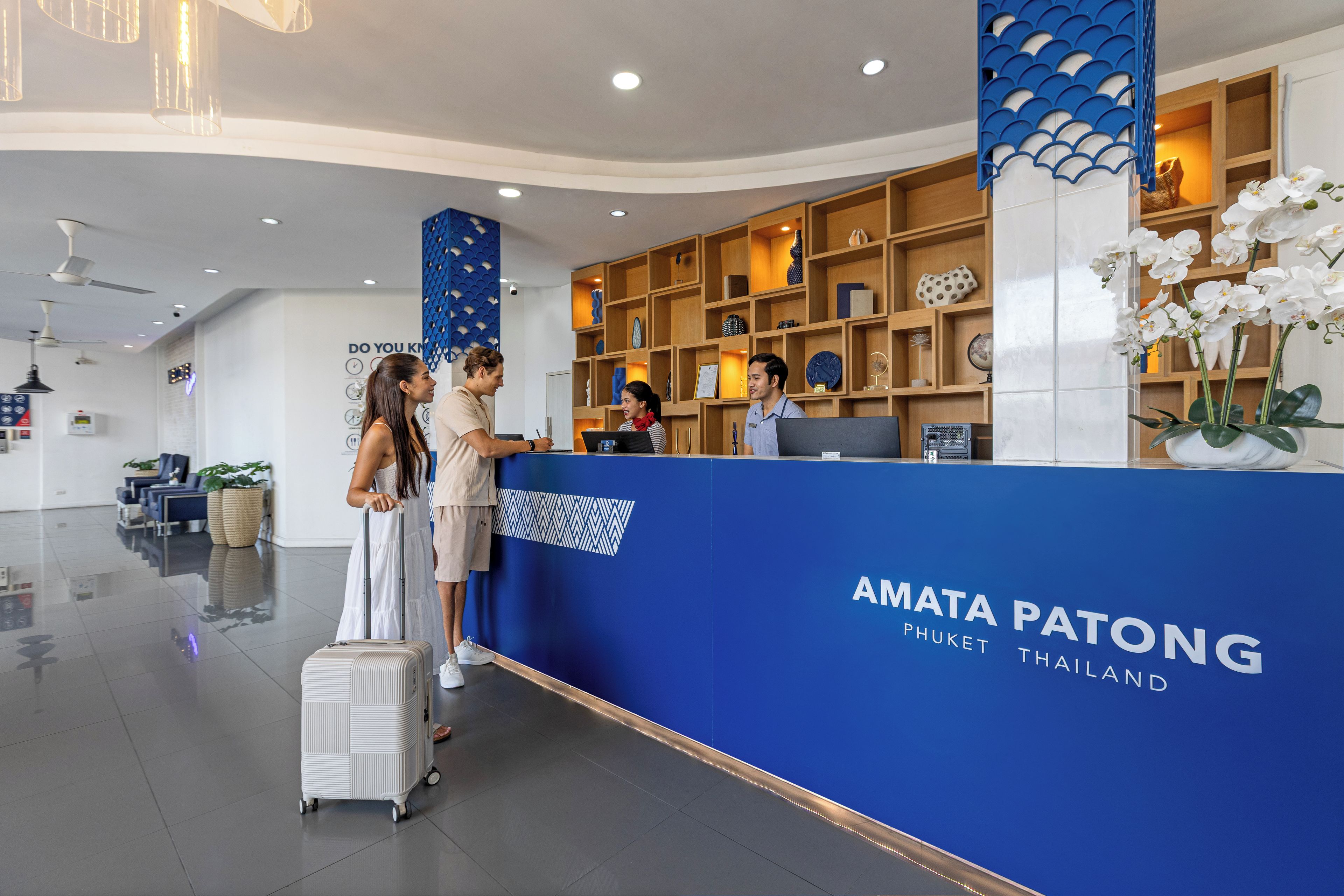 Un mostrador de recepción con una decoración azul y blanca, un cartel que dice 'AMATA PATONG THAILAND' y un área de recepción con un diseño moderno que incluye un ventilador de techo e iluminación decorativa.
