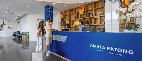 Un mostrador de recepción con una decoración azul y blanca, un cartel que dice 'AMATA PATONG THAILAND' y un área de recepción con un diseño moderno que incluye un ventilador de techo e iluminación decorativa.
