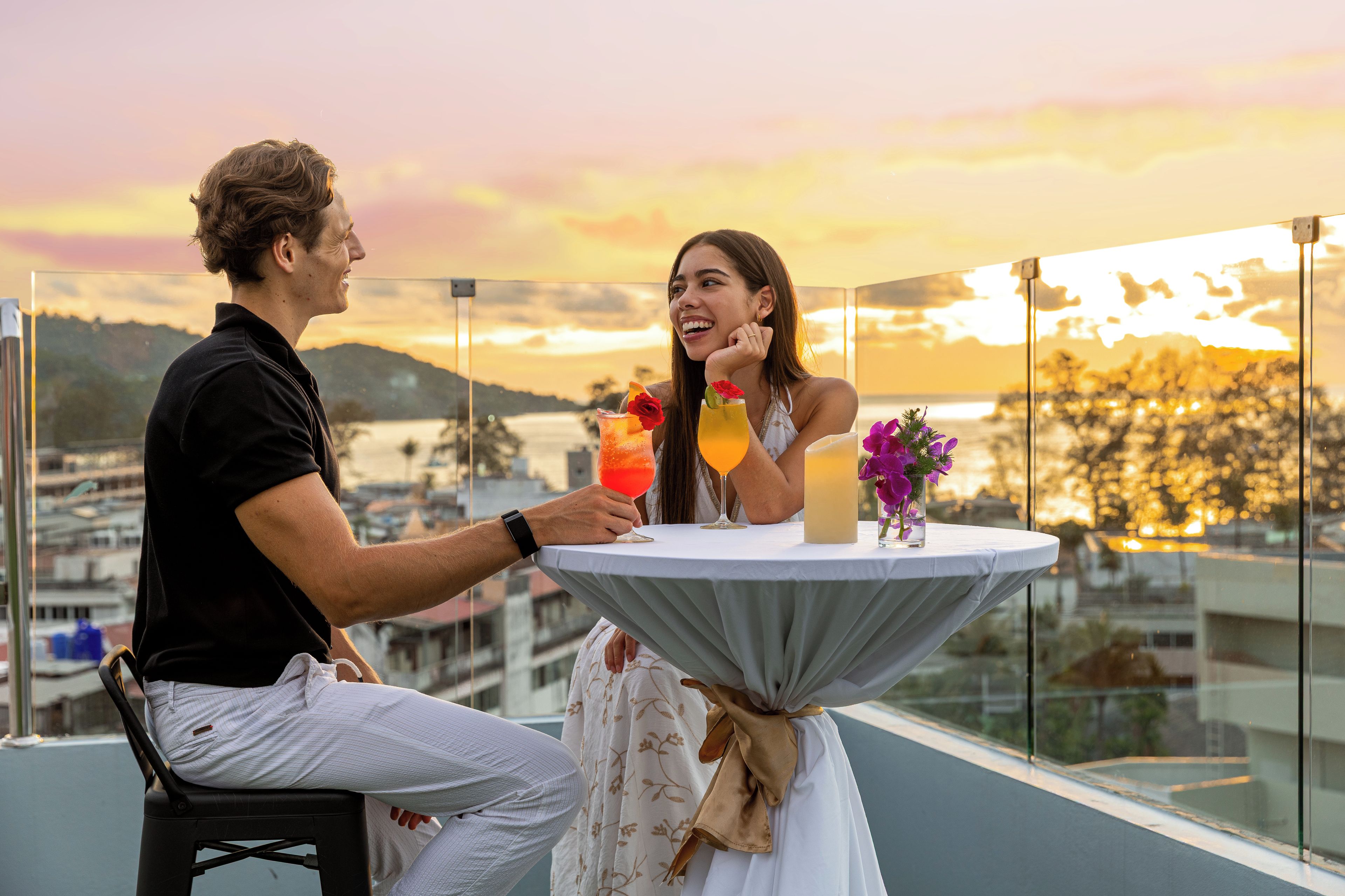 A man and a woman seated at a round table with drinks, overlooking a cityscape at sunset.