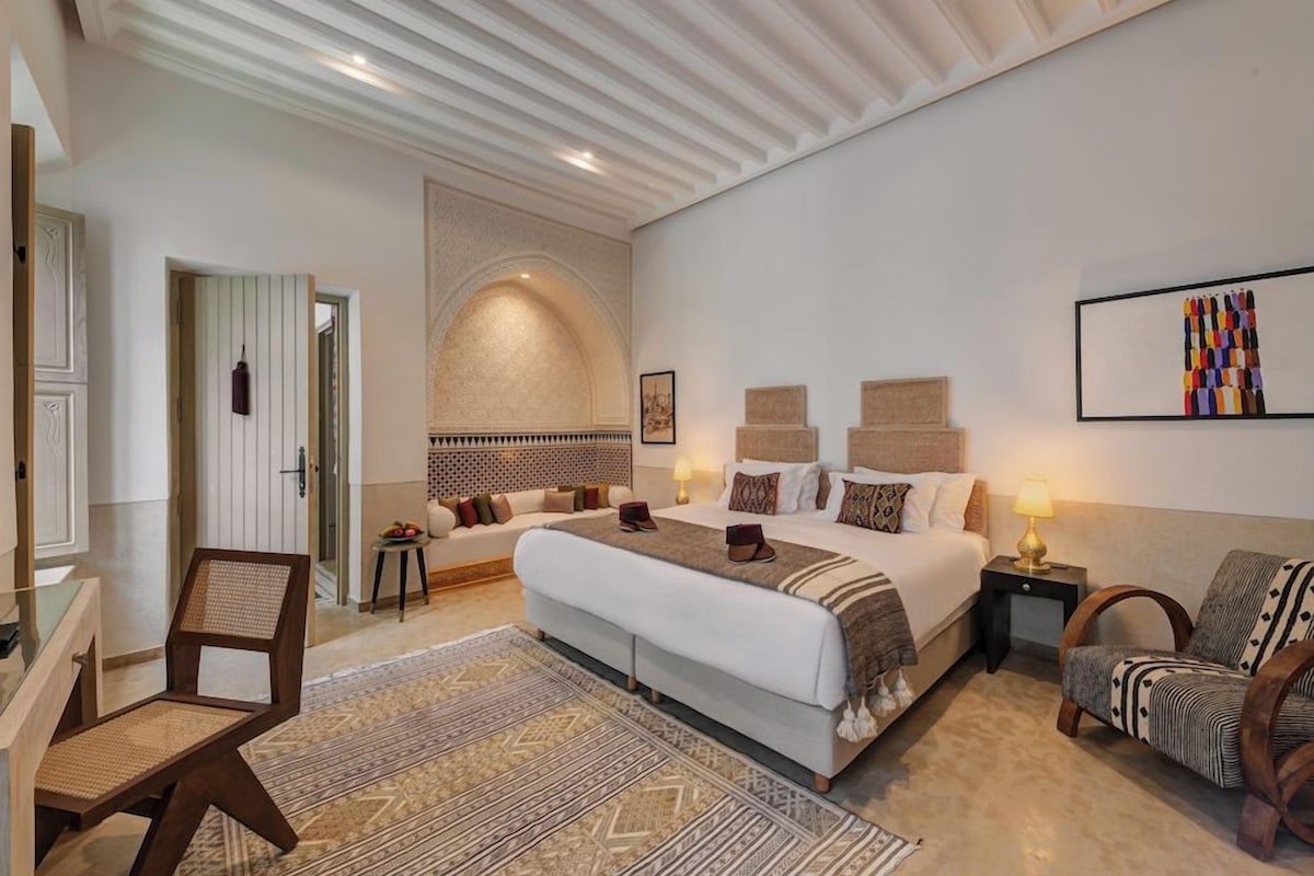 Una habitación de hotel con una cama grande, un área de descanso con un sillón, una mesita con lámpara y un adorno en la pared.