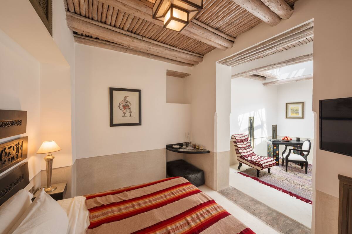 Une chambre à coucher avec un lit, une table de chevet, une lampe, un tableau encadré au mur, une chaise, une table et un téléviseur.