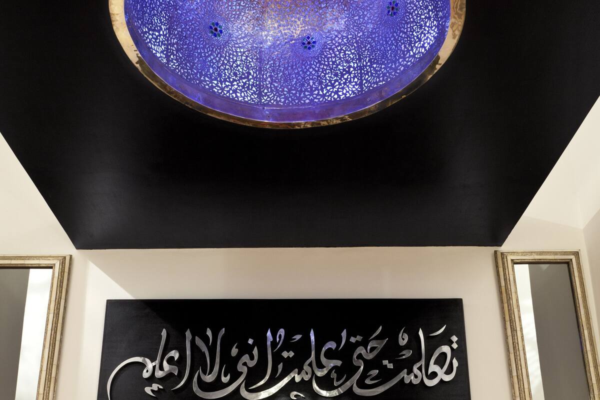 Une chambre à coucher avec un lit, des oreillers, une œuvre d’art murale ornée de calligraphie arabe et un luminaire fixé au plafond.