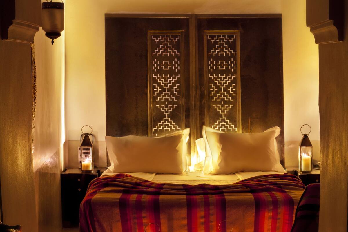 Une chambre  à coucher accueillante avec un lit, des oreillers et un mur décoré d’un motif. On y trouve deux lanternes sur les tables de chevet et une lanterne suspendue au-dessus du lit.