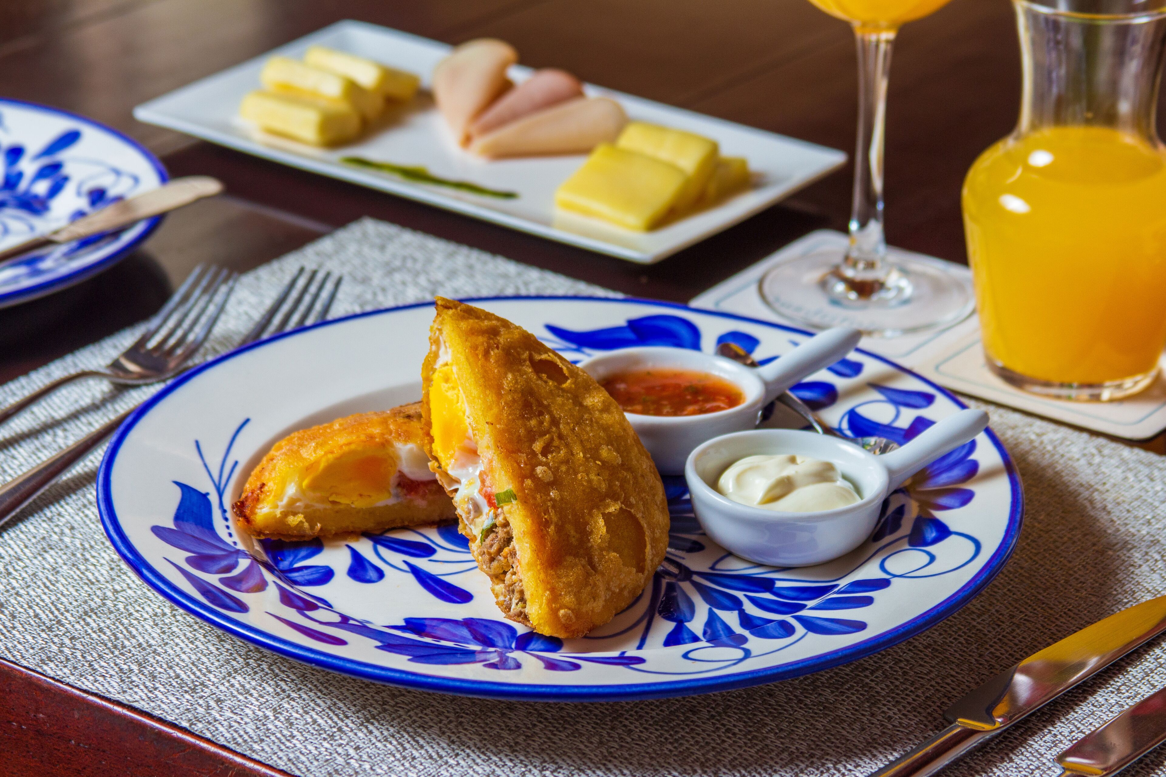 Un plato con un alimento frito, un vaso de jugo de naranja y un pequeño recipiente con salsa.