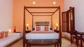 A four-poster bed with a white bedspread and red pillows, a wooden bench in front, and a wooden table with chairs to the right.