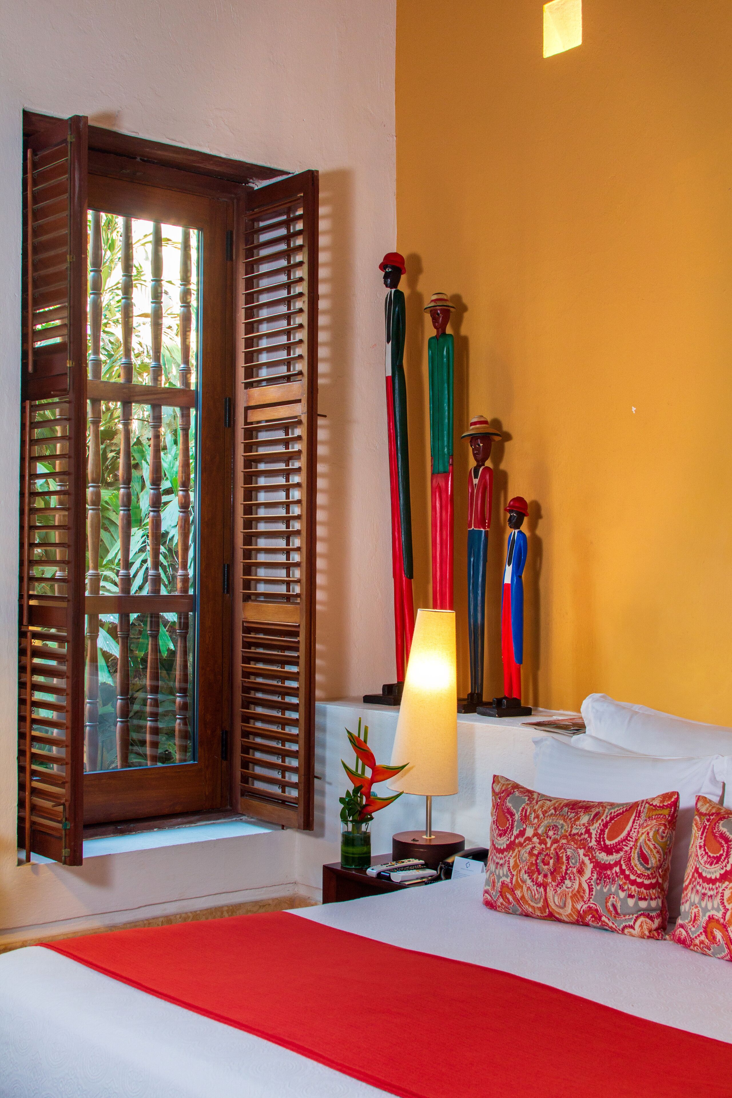 A hotel room with a bed, a window with wooden shutters, a nightstand with a lamp, and decorative items.