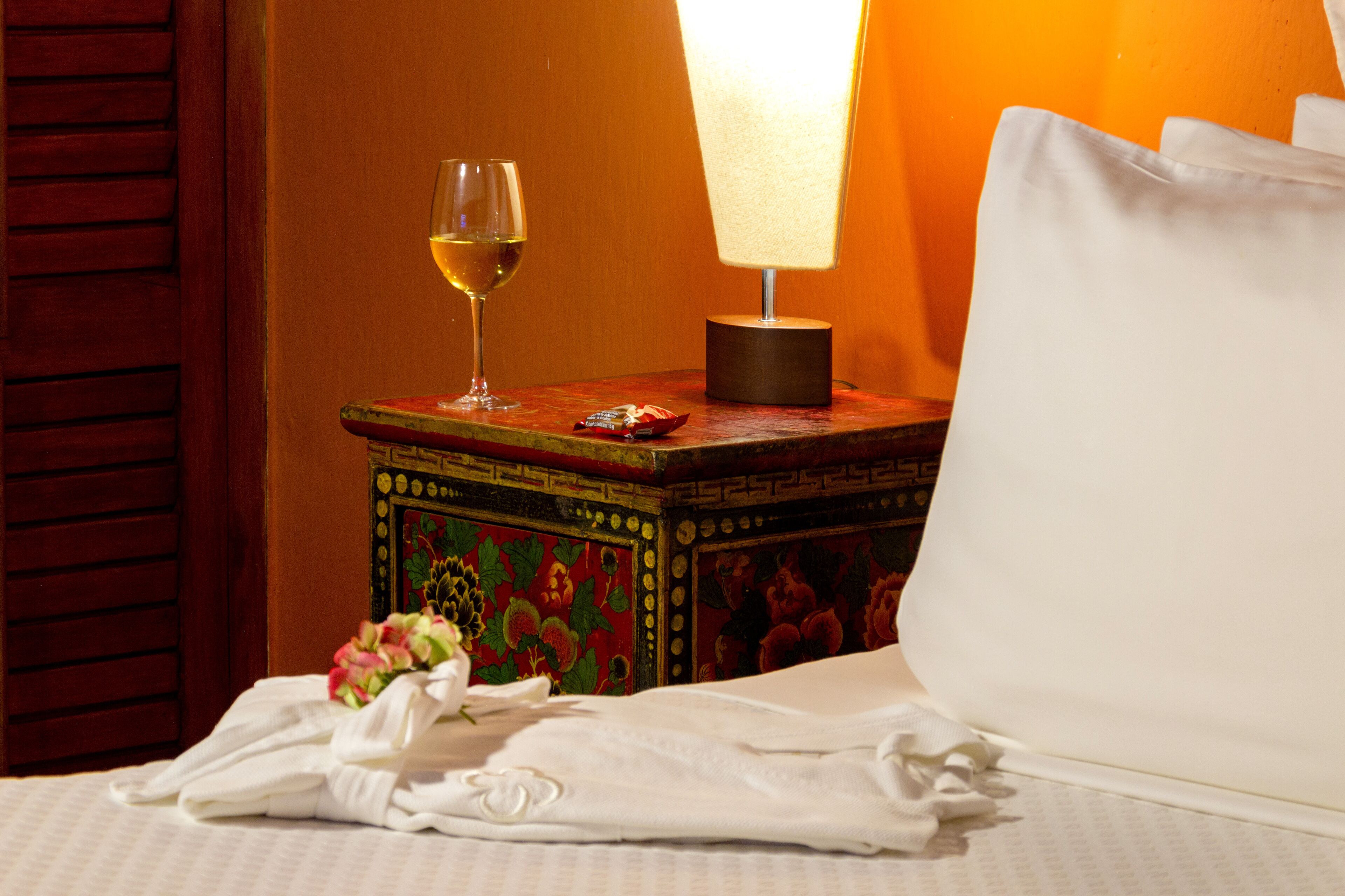 A neatly made bed with a white pillow, a glass of white wine, a small lamp, and a decorative box on a wooden nightstand.