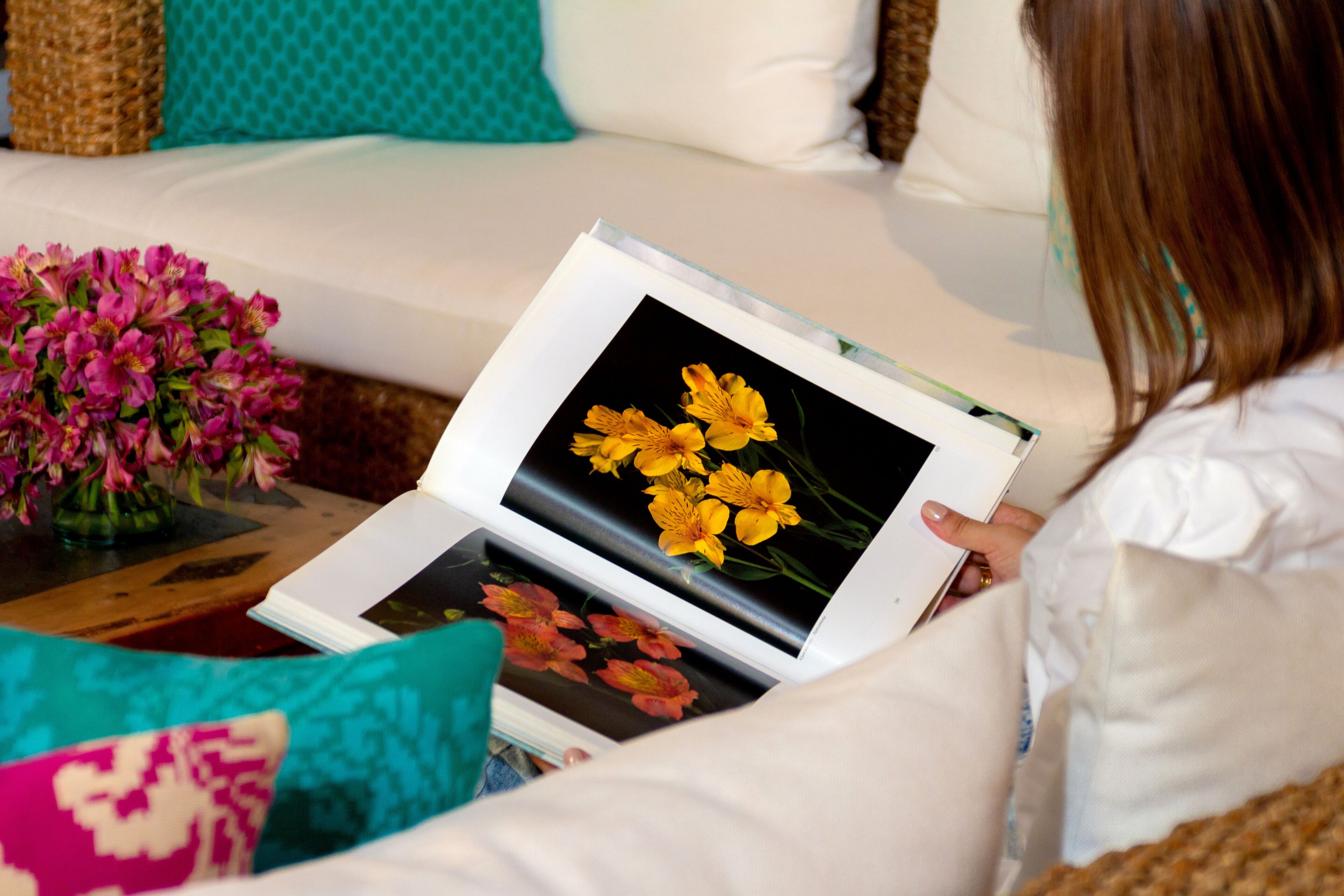 A person sitting on a white couch with teal cushions, holding a book with a picture of yellow flowers on the cover.