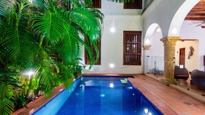 An indoor swimming pool with tiled walls and a wooden floor, surrounded by lush green plants and a white building with wooden window frames. - Hotel Quadrifolio (Cartagena)