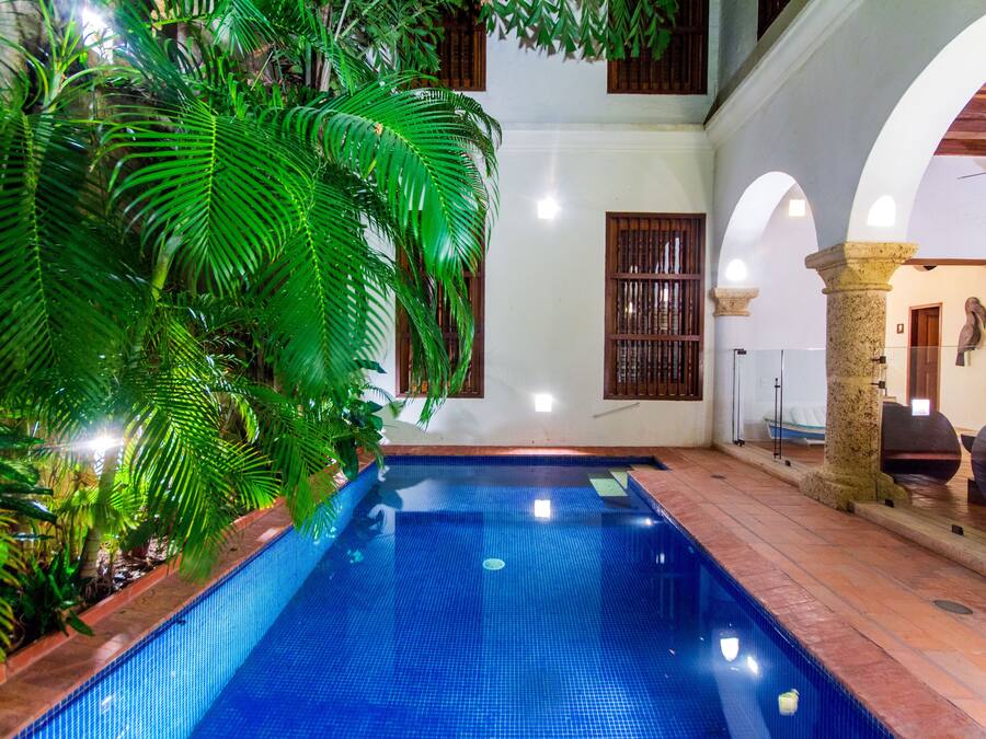 An indoor swimming pool with tiled walls and a wooden floor, surrounded by lush green plants and a white building with wooden window frames.