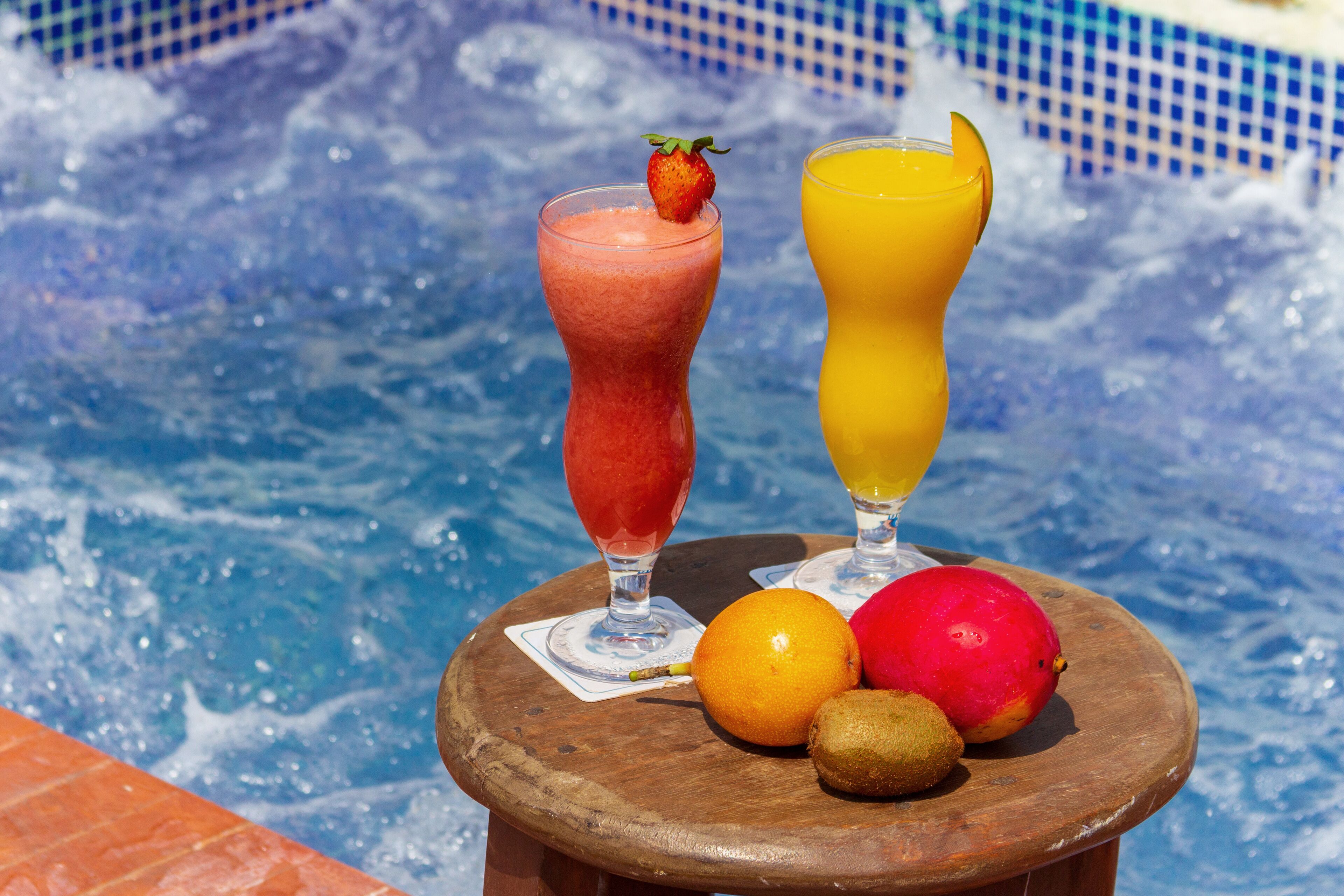 A wooden table with two drinks and fruits by a pool.