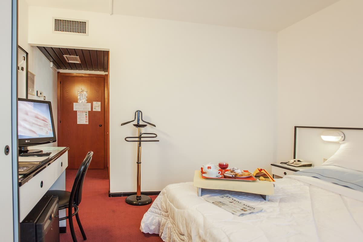 A hotel room with a bed, a desk with a computer, a chair, a bedside table with a lamp, and a door with a sign.