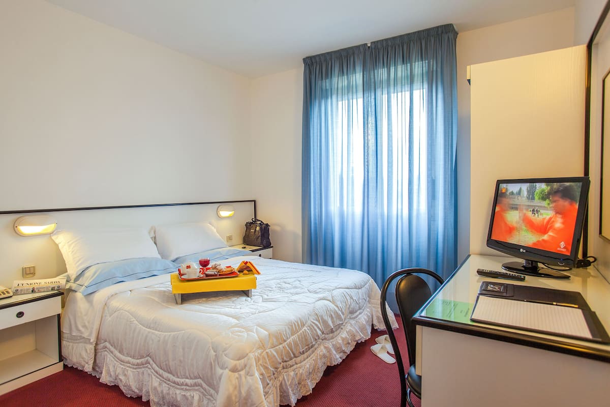 A hotel room with a bed, a desk with a computer, a chair, a nightstand, and a window with curtains.