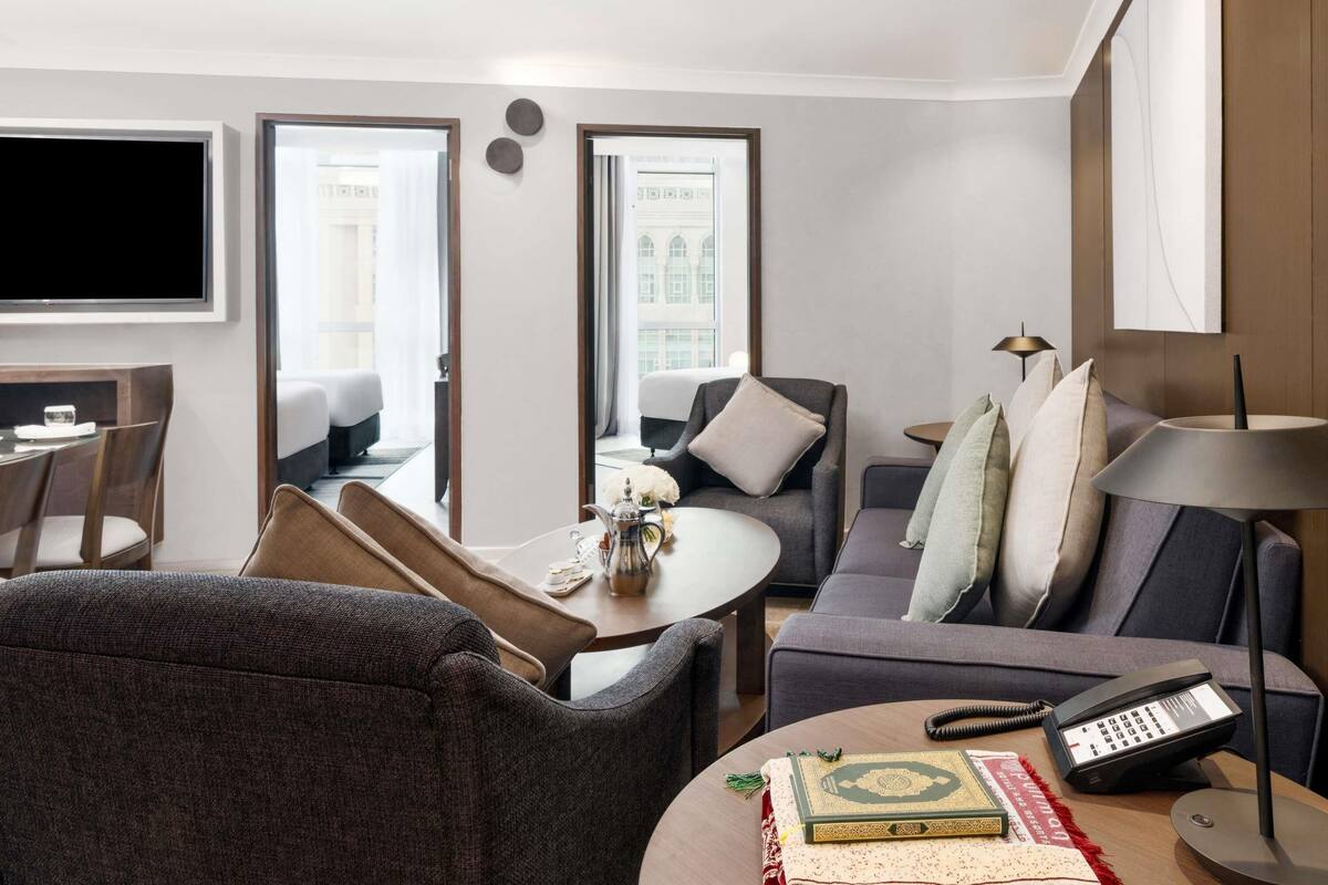 A modern hotel room with a sofa, armchair, dining table, and a TV.