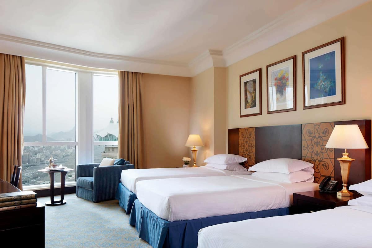 A hotel room with two beds, a desk, a chair, and a view of a cityscape through large windows.