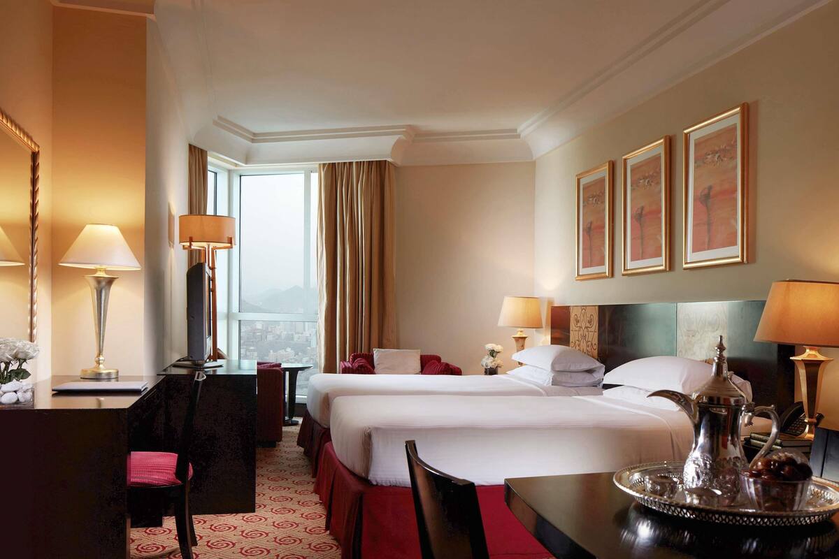 A hotel room with two beds, a desk, a chair, a dining table, and a view of the city.