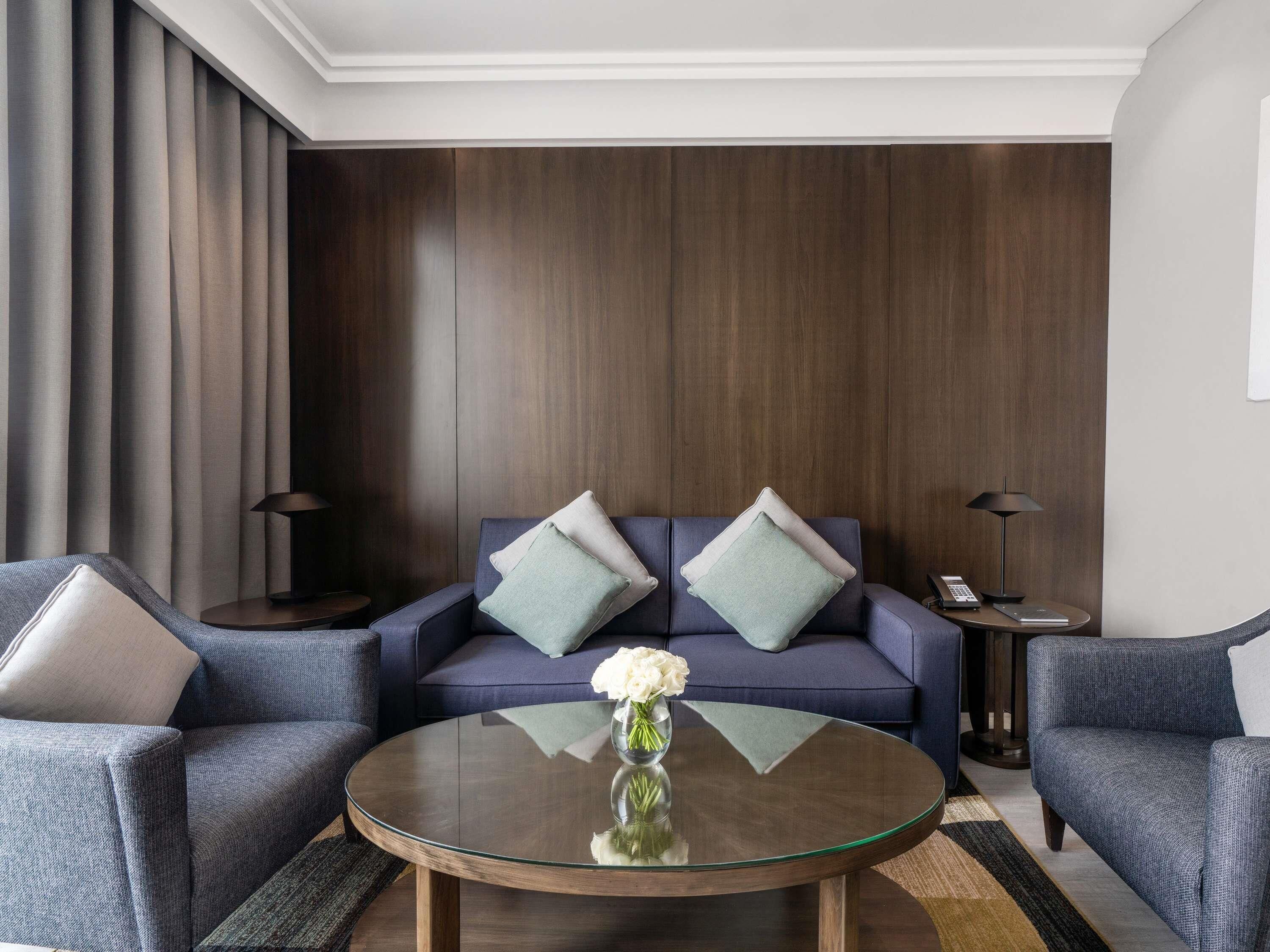 Una sala de estar moderna con un revestimiento de madera oscura, un sofá azul con dos cojines, una mesa de centro de vidrio con un jarrón de flores y dos sillones.