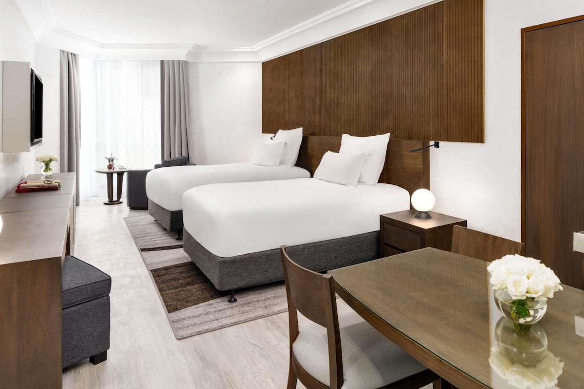 A hotel room with two beds, a desk, a chair, a small table, and a vase of flowers.