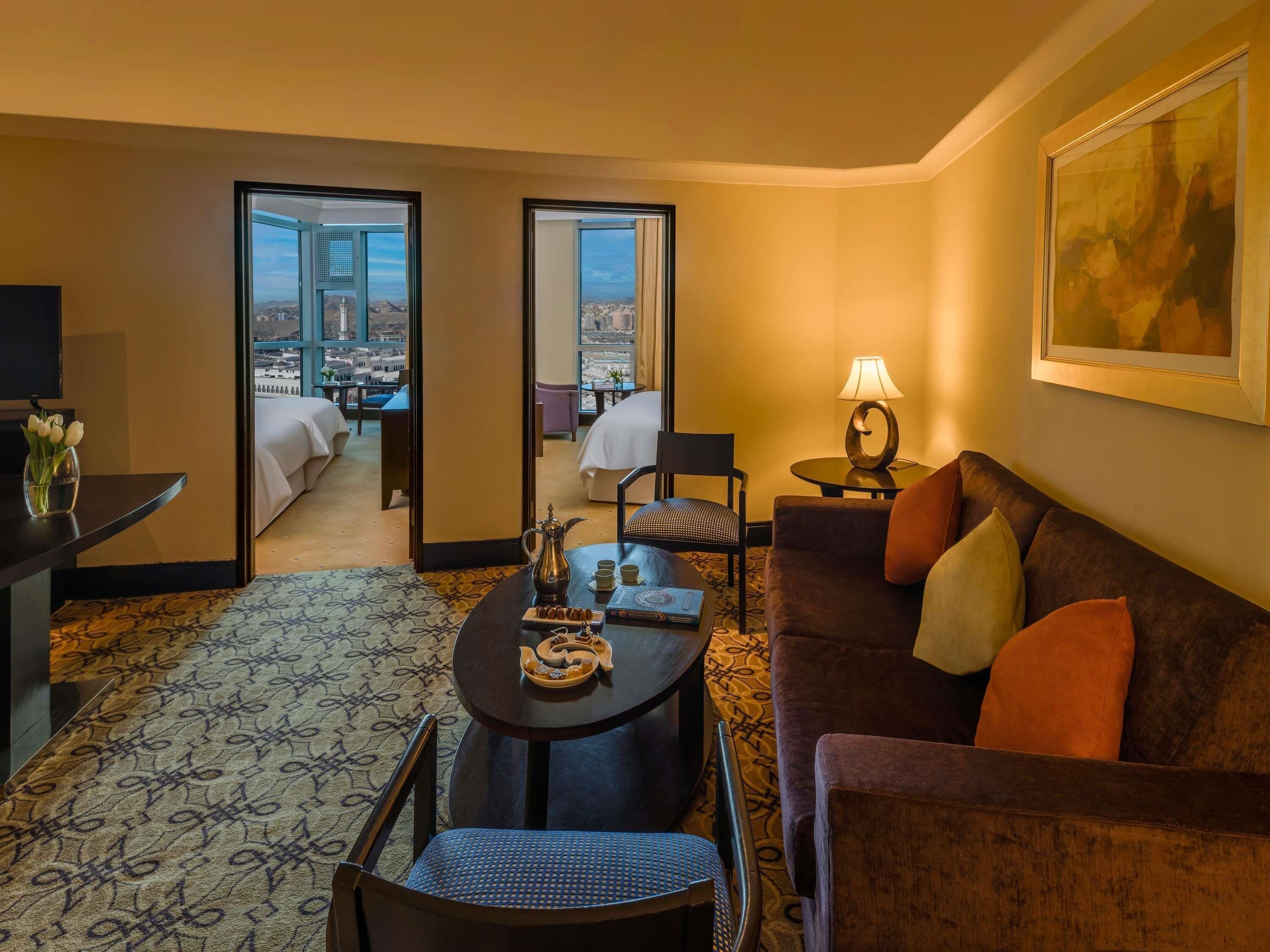 A hotel room with a dining area, a sofa, a lamp, a painting, and a view of a cityscape through large windows.