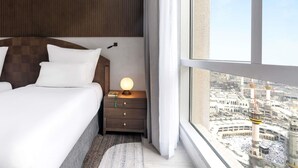 A hotel room with a large bed, a nightstand with a lamp, and a view of a cityscape through a large window. - Zamzam Pullman Makkah (Makkah)
