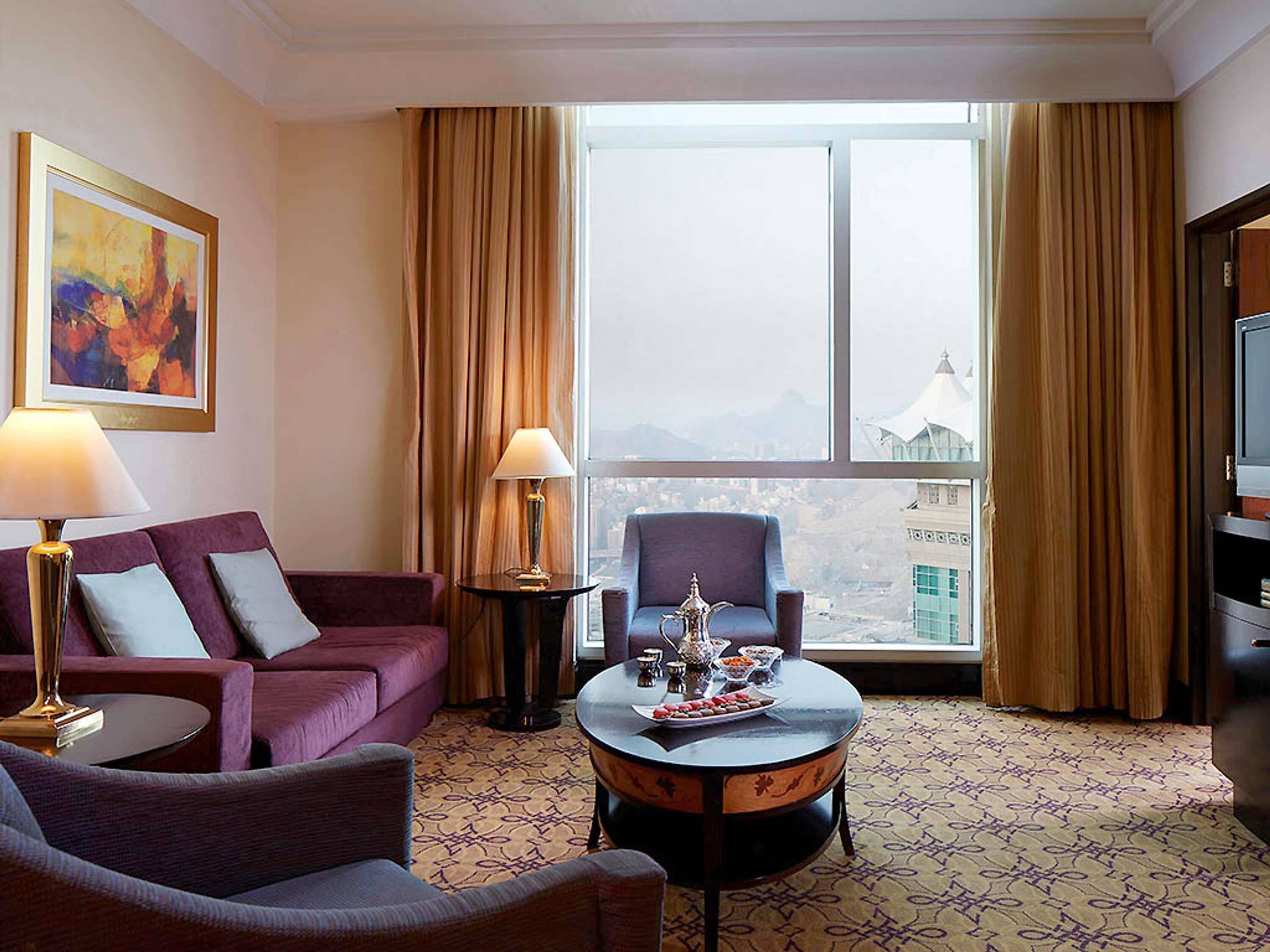 A hotel room with a sofa, armchair, coffee table, lamp, and a television.