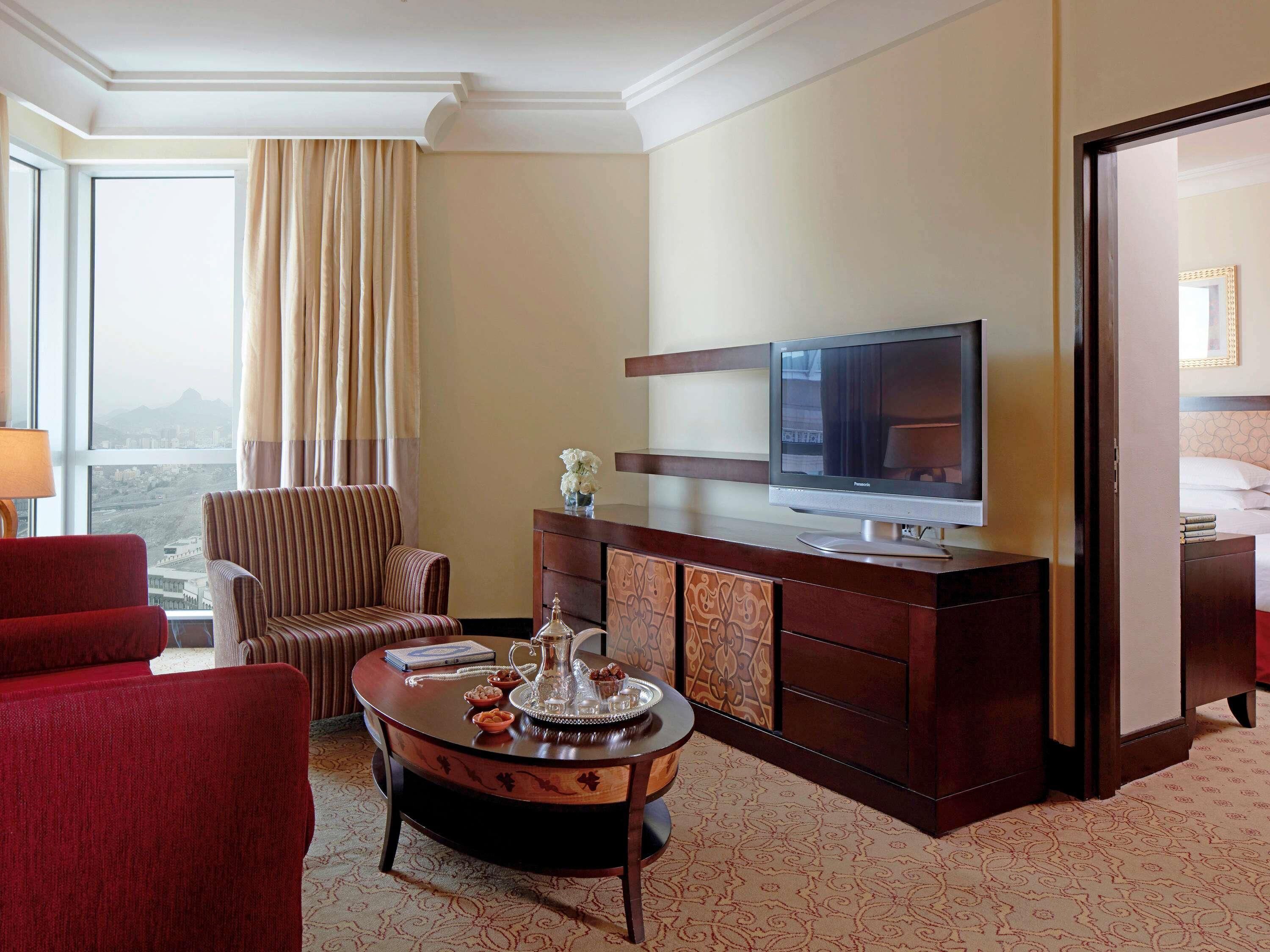 Une chambre d’hôtel dotée d’une grande fenêtre, d’un téléviseur, d’un fauteuil rouge, d’une chaise à rayures, d’une table basse en bois et d’un mini-bar.