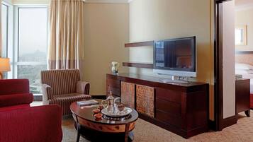 Une chambre d’hôtel dotée d’une grande fenêtre, d’un téléviseur, d’un fauteuil rouge, d’une chaise à rayures, d’une table basse en bois et d’un mini-bar.