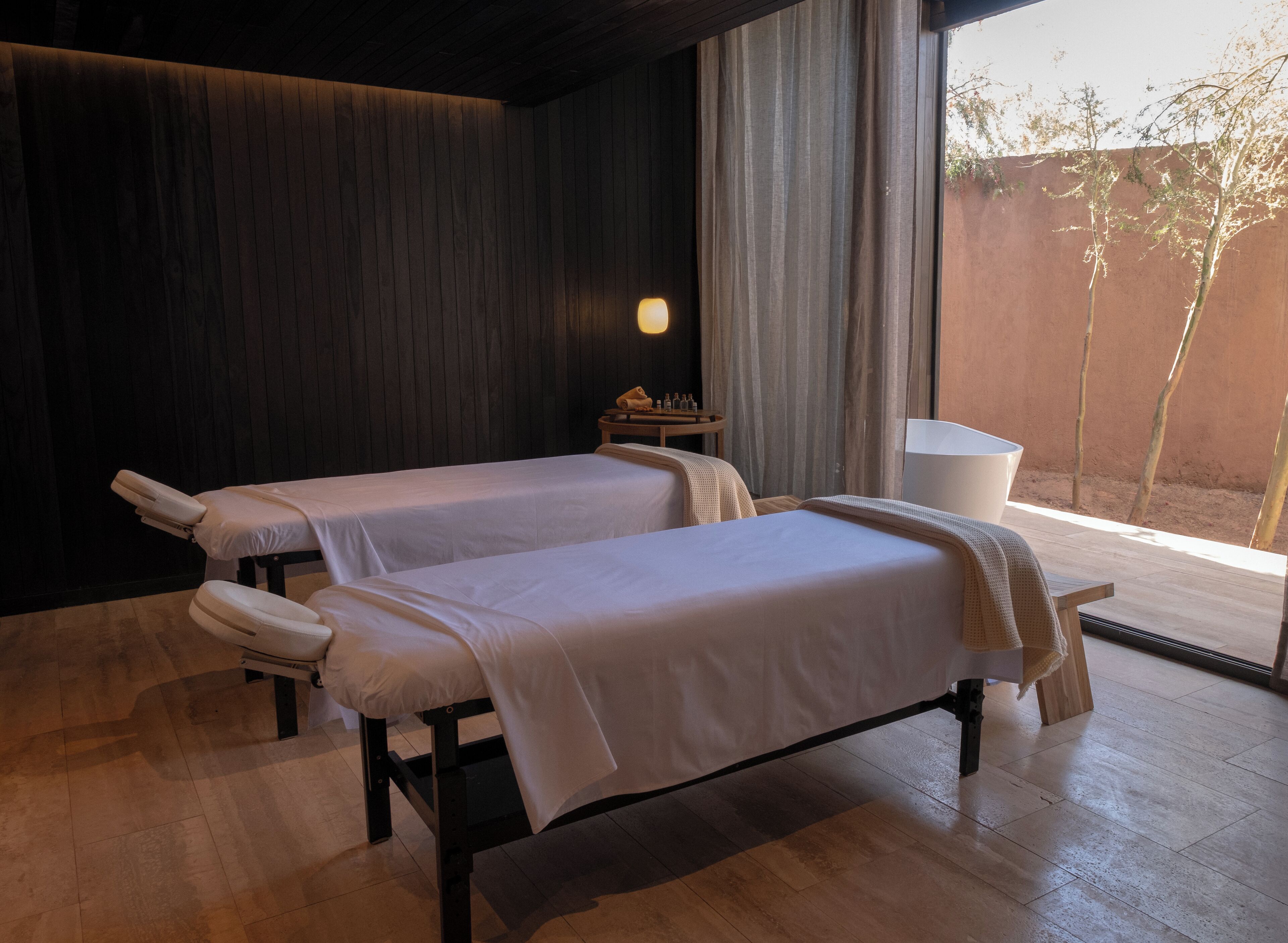Un cuarto de spa con dos camillas, una mesita con botellas y vistas a árboles a través de una ventana.